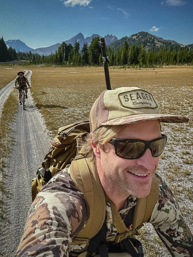A man taking a selfie while hiking in a remote, mountainous area with a wide trail. He is wearing sunglasses, a cap, camouflage shirt, and a backpack, with a woman riding a bike on the trail in the background. The scenery includes trees and distant m