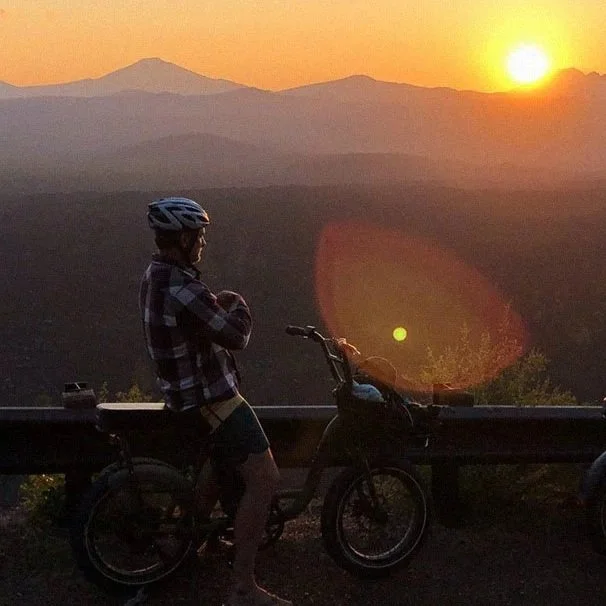 Person wearing a helmet and checkered shirt standing next to a bicycle at sunset with mountains in the background.