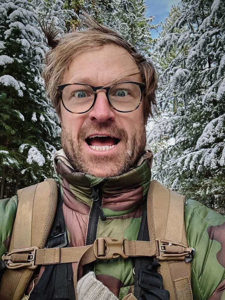 A man with glasses taking a selfie outdoors in a snowy forest, wearing a camouflage jacket and a backpack.