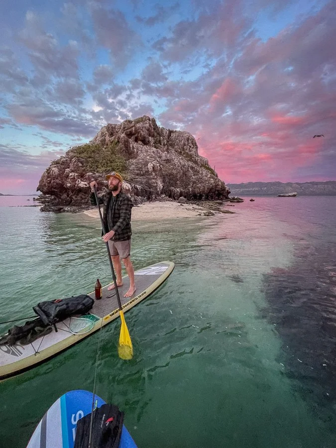 Man paddleboarding on calm water near a rocky island during sunset, with colorful sky and clouds.