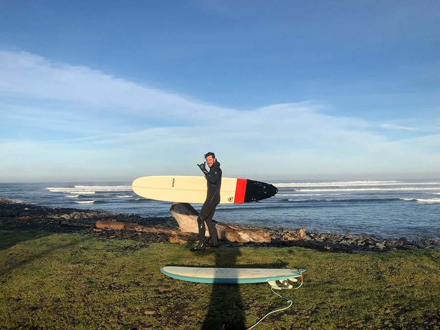 Man in a wetsuit holding a surfboard near the shoreline with an ocean and blue sky in the background.