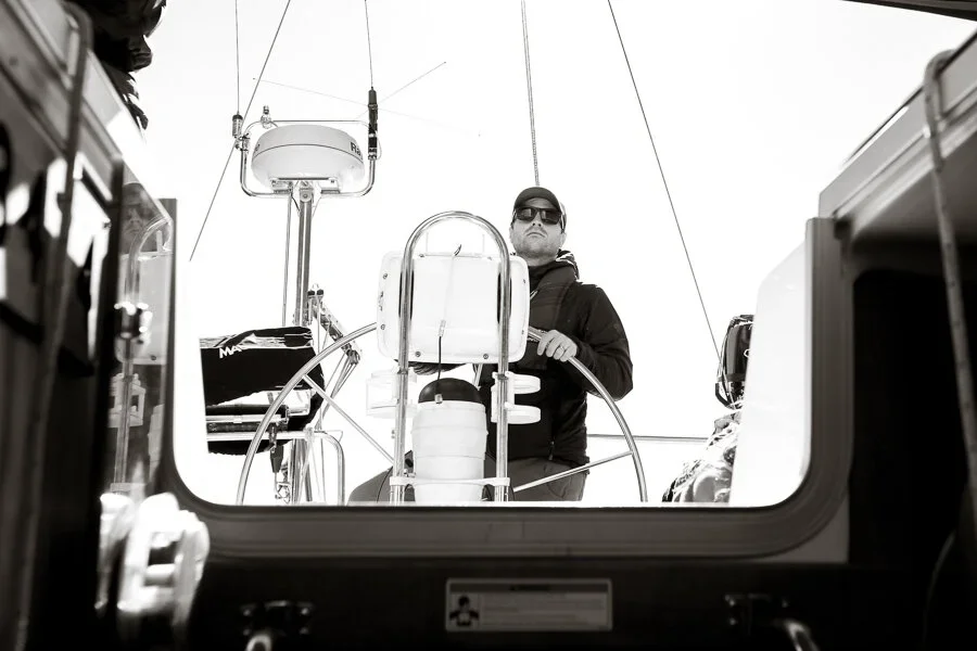 View through a boat hatch showing a man at the helm, wearing sunglasses and a cap, with sailing equipment and rigging around him.
