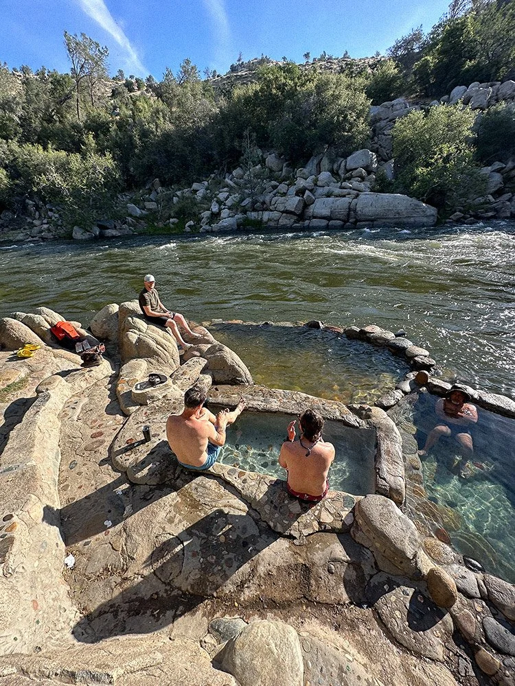People relaxing in hot tubs near a river in a rocky, forested outdoor setting