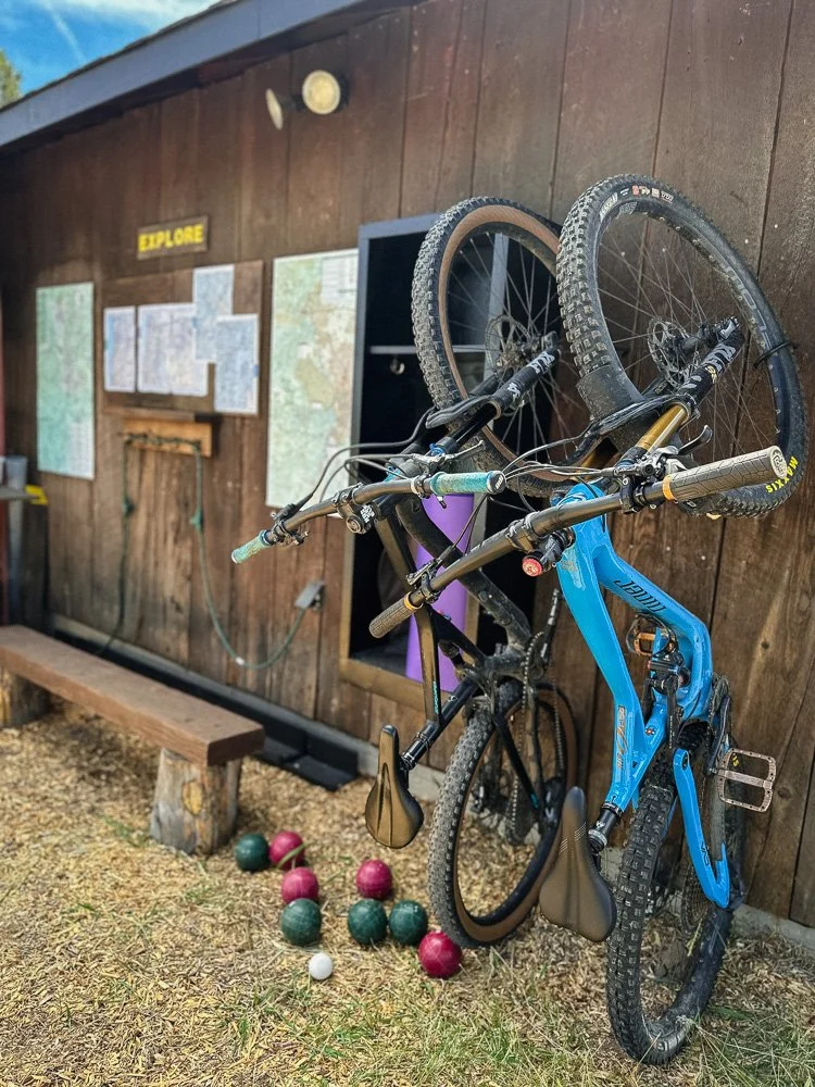 A blue mountain bike hanging on a wooden wall at outdoor adventure site with a sign that says 'Explore', maps, and a small bench nearby. Several colorful balls are on the ground in front of the wall.