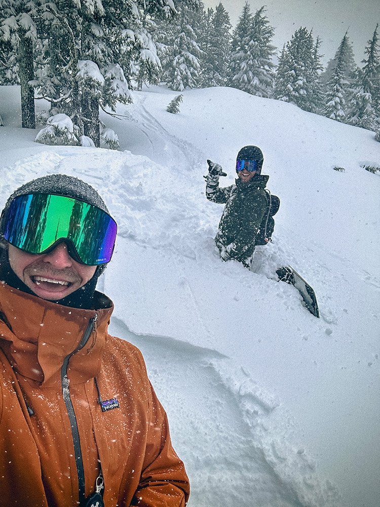 Two people in winter gear enjoying a snowy landscape. One in the foreground wearing an orange jacket and reflective goggles, smiling at the camera. The other in the background standing in deep snow, wearing a black jacket, helmet, and goggles, giving