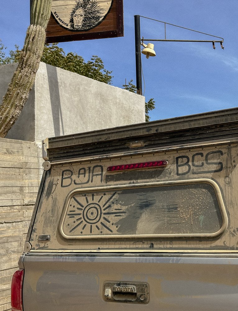 The back of a weathered Toyota truck with graffiti that reads 'BAJA BCG,' a sun drawing on the rear window, and a red brake light strip. The background includes a gray concrete wall, a cactus, a billboard with a face illustration, and a blue sky with