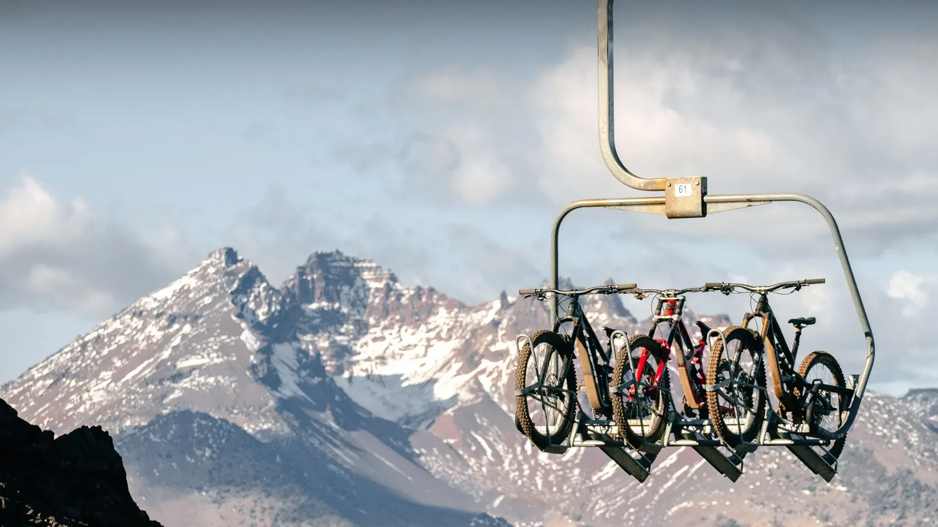 Four mountain bikes hanging from a metal ski lift structure against a background of snow-capped mountain peaks under partly cloudy sky.