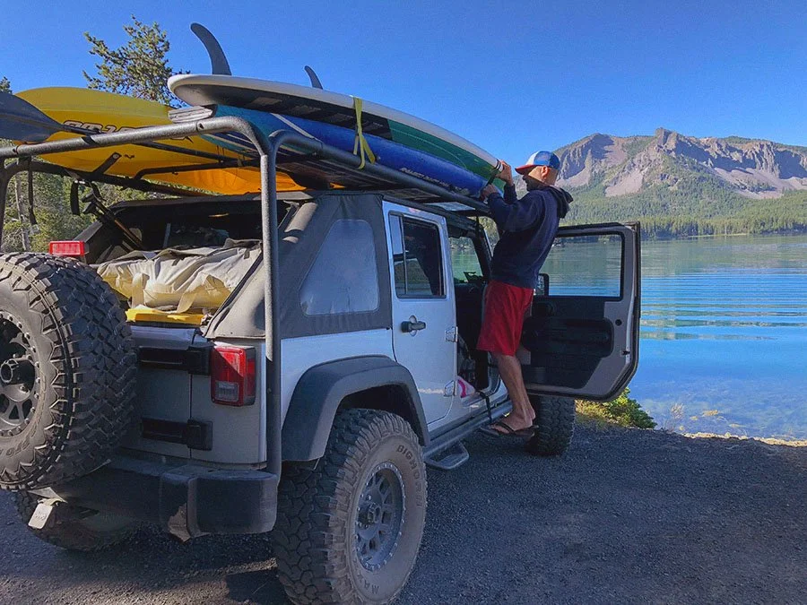 A man preparing a kayak on top of a gray Jeep parked by a lake with mountains in the background.