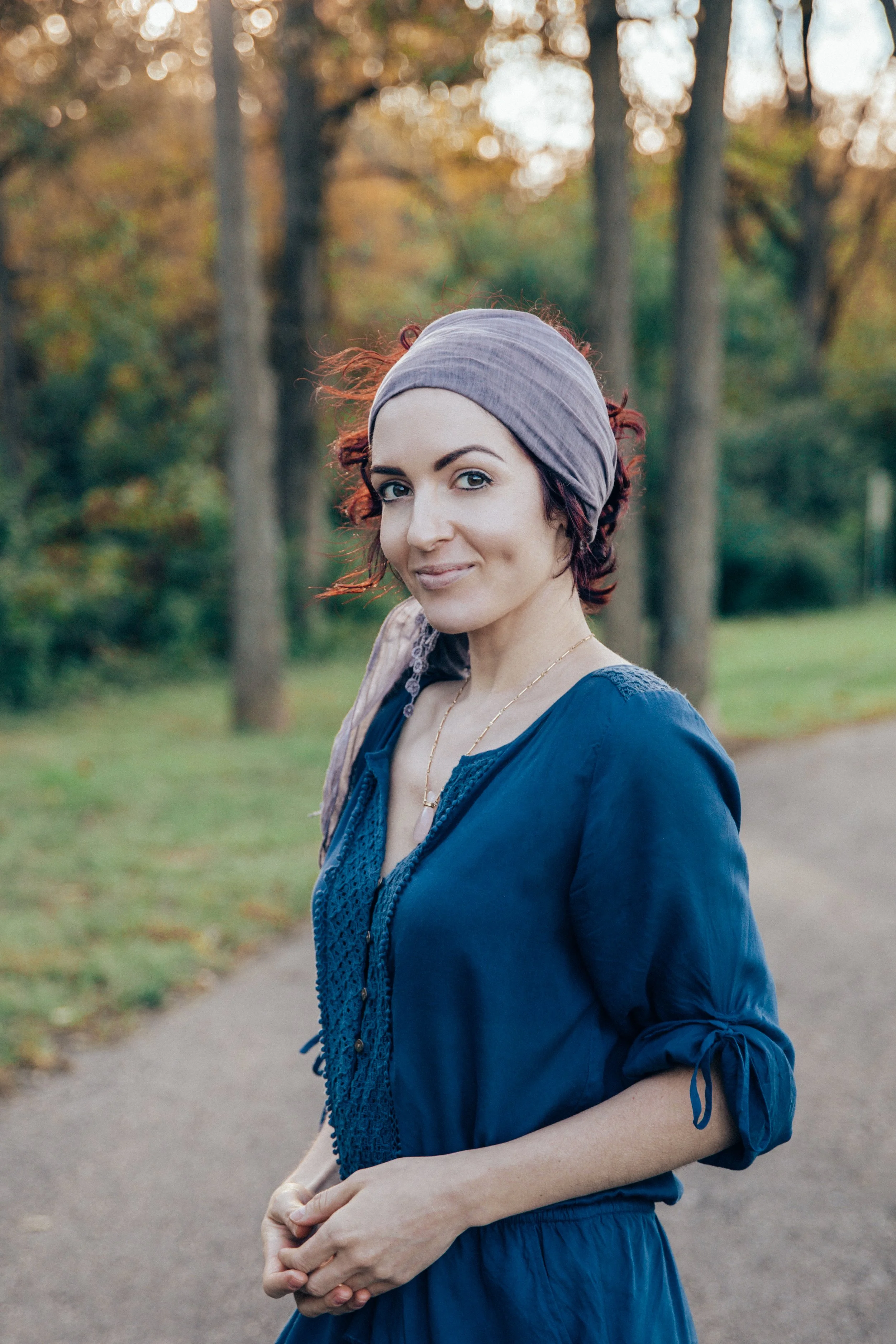 Tara, a white woman with brown eyes, has her dark red hair pulled back by a long purple scarf. She's wearing a deep blue outfit, standing on a path before trees with autumn colors beginning to show.