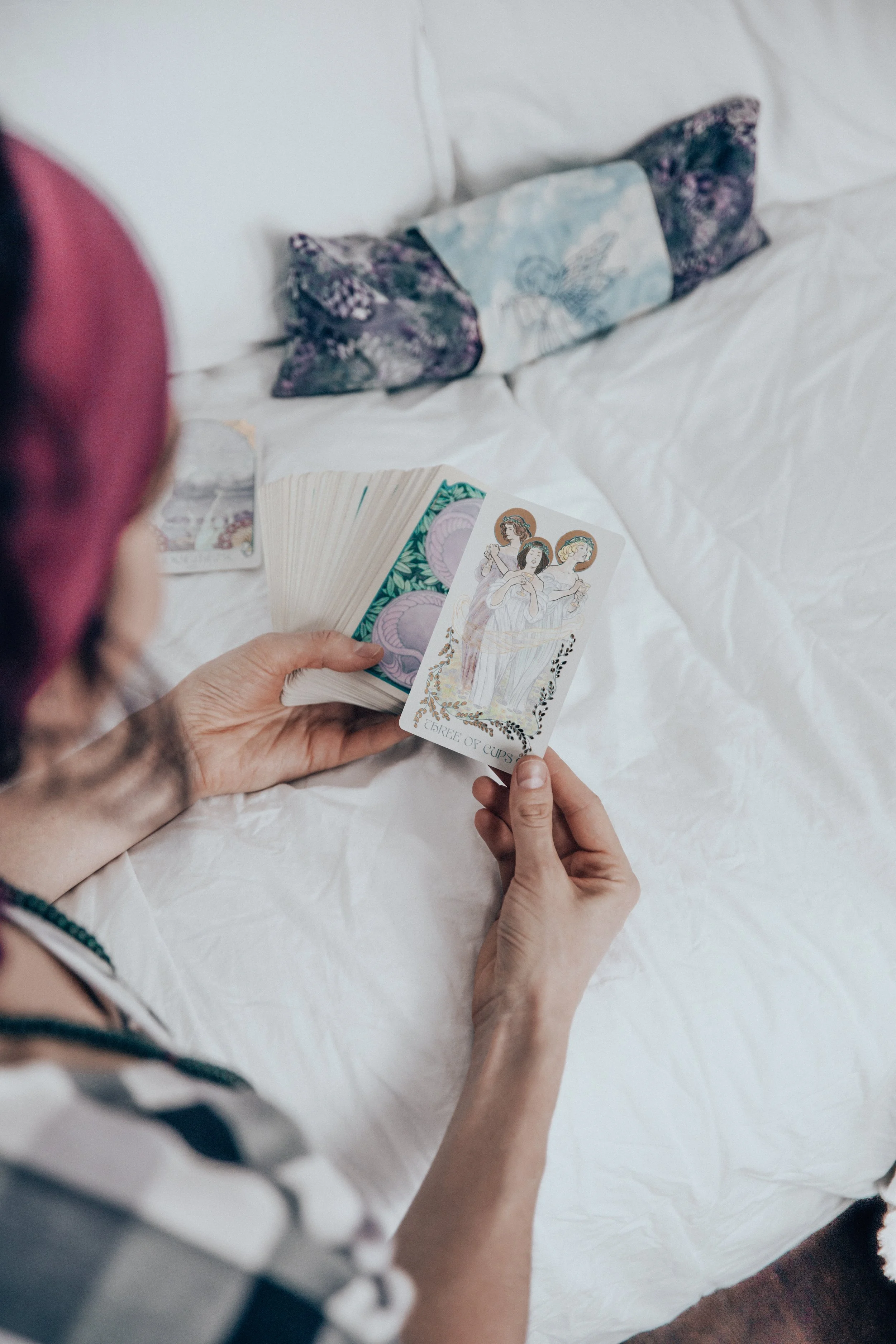 The viewer is placed as though looking over Tara's shoulder with her. She holds a deck of tarot cards, showing us a card with three women with halos around their heads, standing in a circle together as if singing.