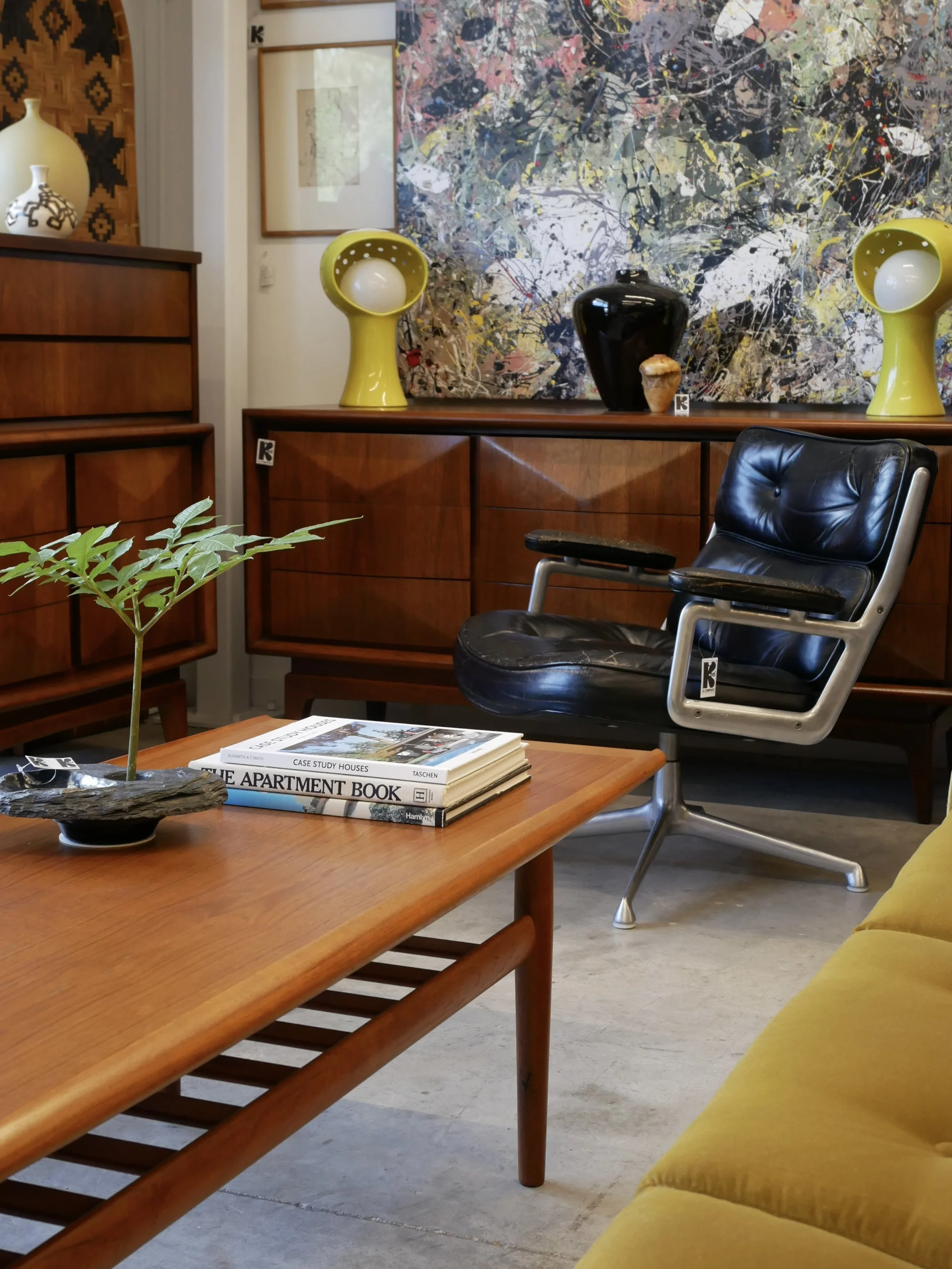 Interior of a modern living room featuring a wooden coffee table with a small plant and magazines, a black vintage-style office chair, a yellow velvet sofa, and a wooden sideboard with colorful vases and an abstract painting in the background.
