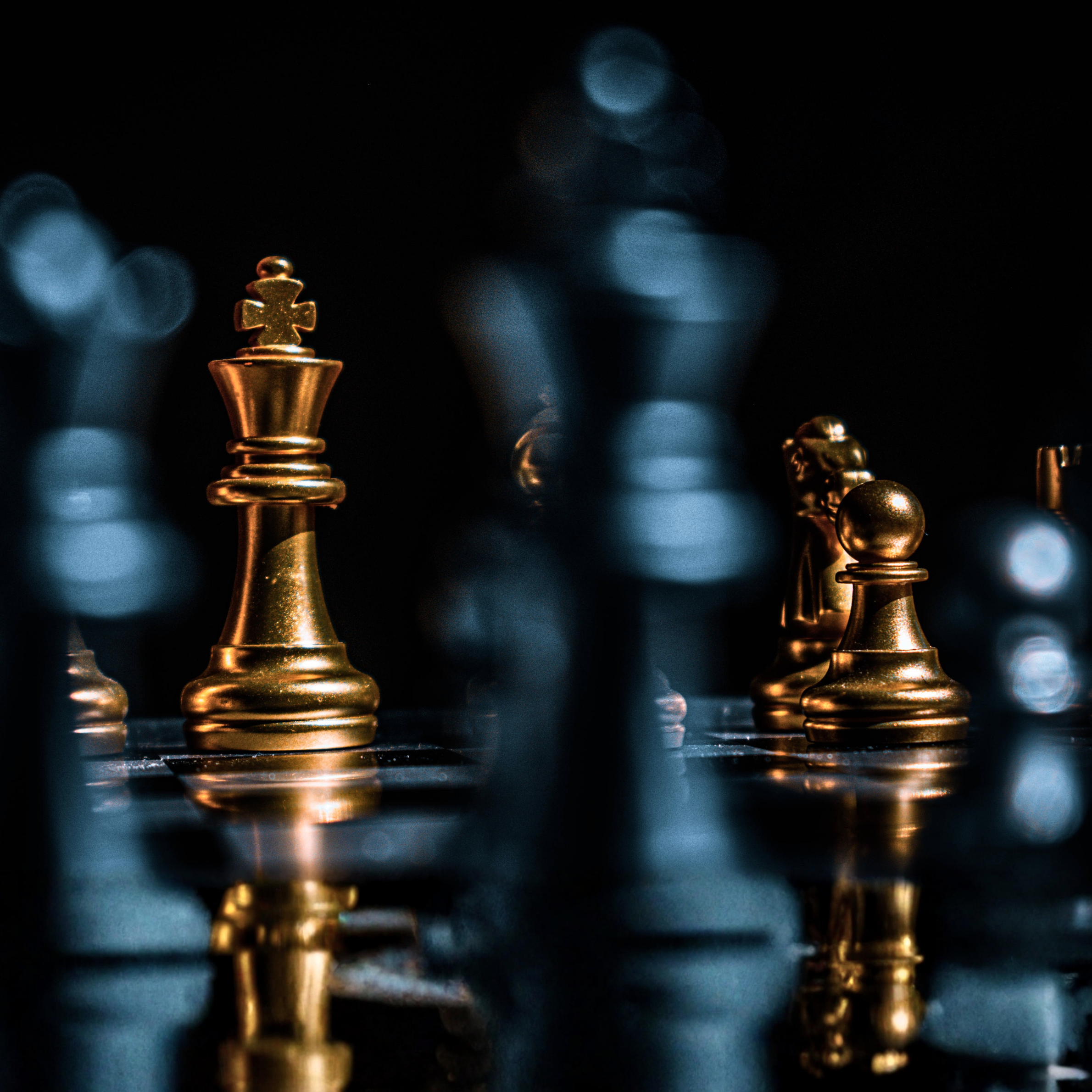 Closeup of chess pieces showing a king and two pawns in metallic gold against a deep gray background with scattered blue boka lights.