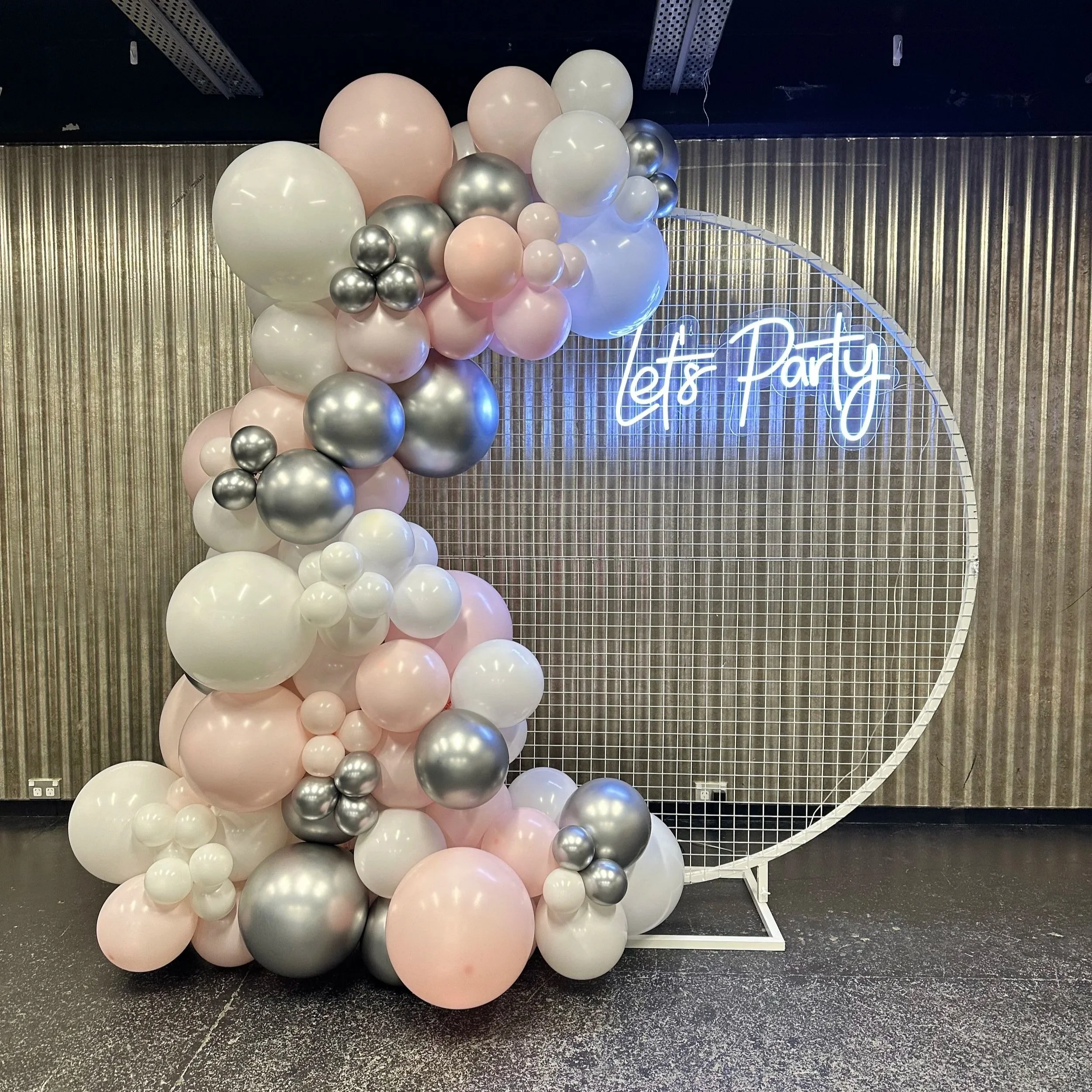 Decorative balloon arrangement with white, metallic silver & gold balloons next to a circular wire grid backdrop with a neon sign reading 'Twenty One'.
