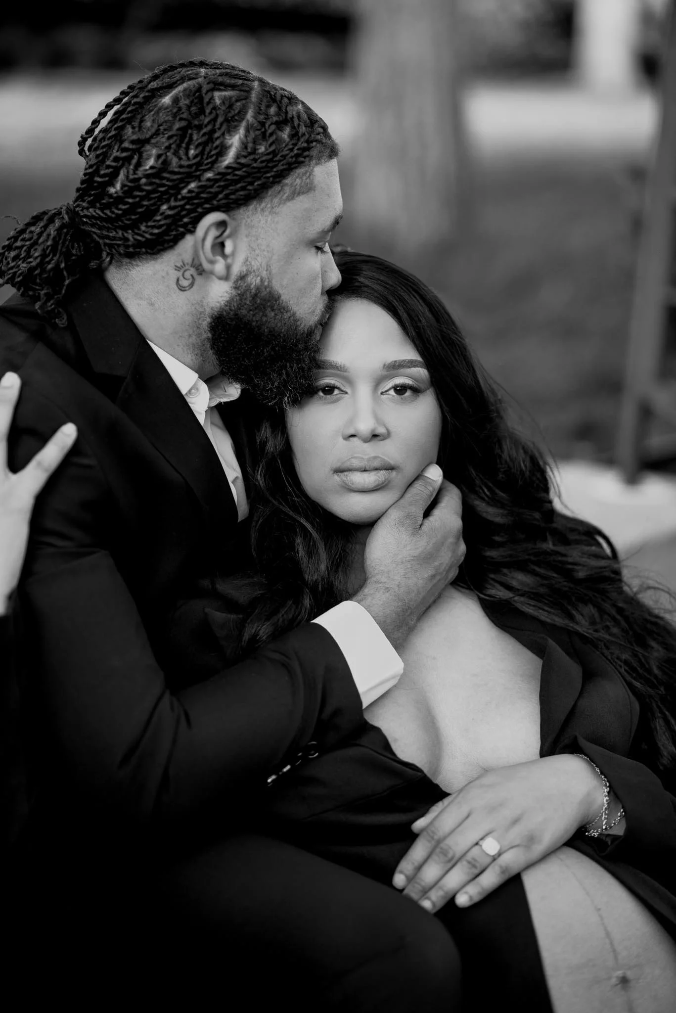 A man with braids and a beard gently holds a woman's face while kissing her on the forehead outdoors. The woman has long dark hair and a neutral expression, wearing a partially open jacket revealing her shoulder.