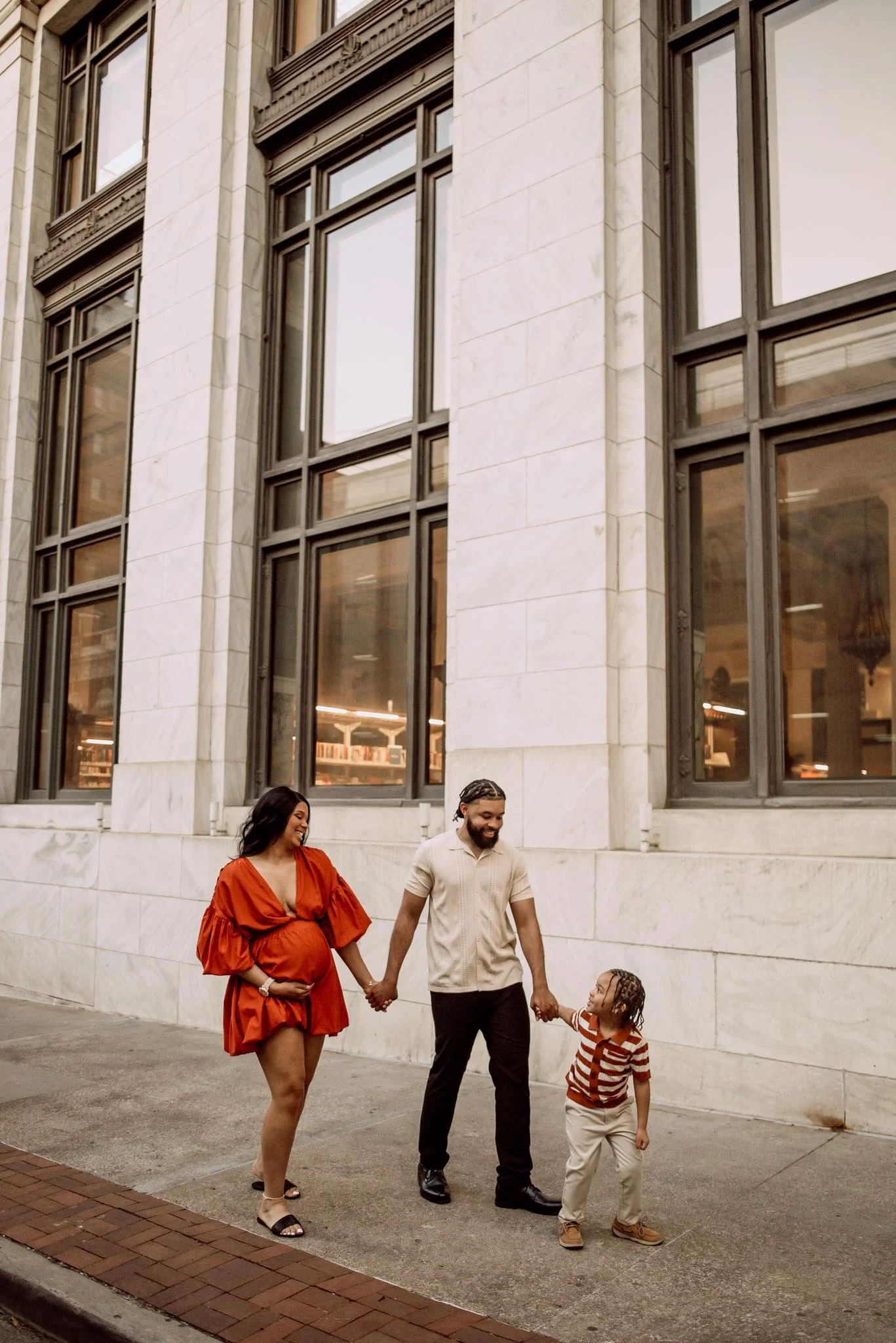 A family walking hand in hand outside a large stone building. The woman is pregnant and wearing a red dress, the man is in a beige shirt, and a young girl in striped shirt and beige pants.
