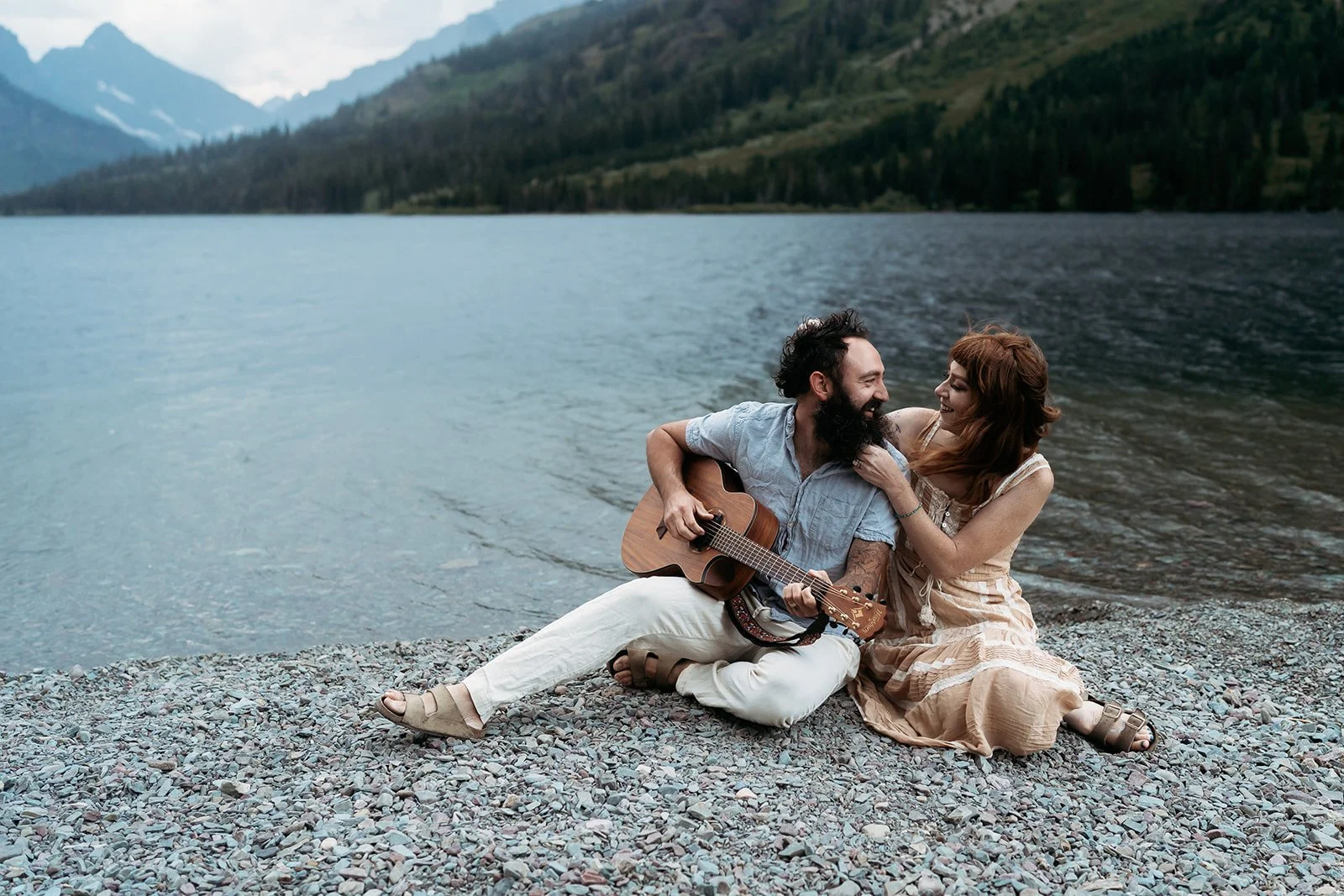 Glacier National Park Elopement photographer captures couple playing guitar and snuggling near two medicine lake, Montana. couple is wearing brown dress and blue linen outfit