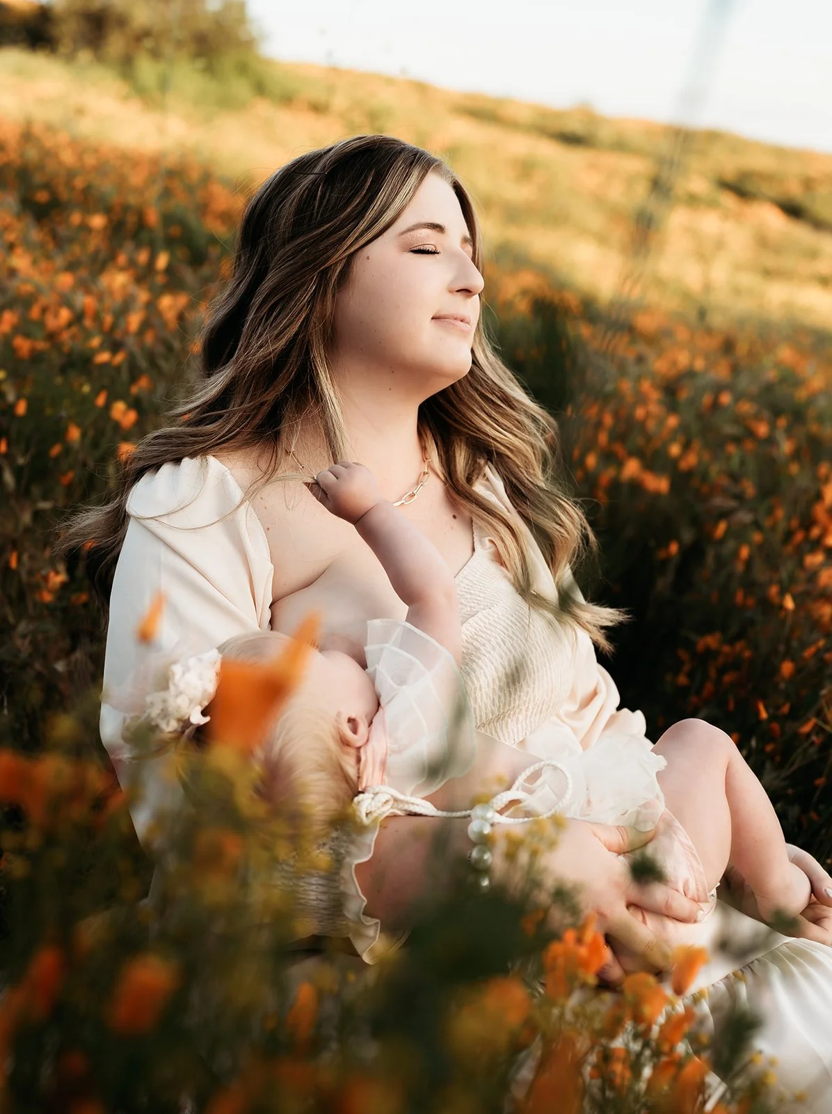 Southern California family photographer captures mother and daughter in a poppy covered field in Riverside California. Mother breastfeeds her daughter and looks out to the sunset