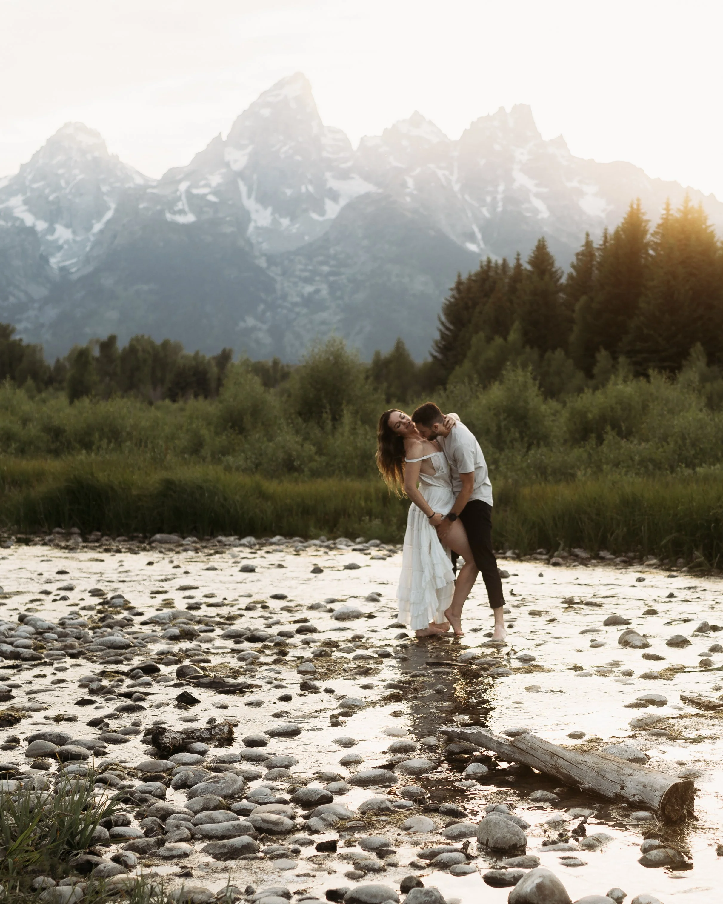 grand Teton national park elopement photographer captures couple near Schwabacher's Landing snuggling with the Teton's in the background. Bride is wearing a white dress and groom is wearing tan bylt shirt in this moody, rich edit.
