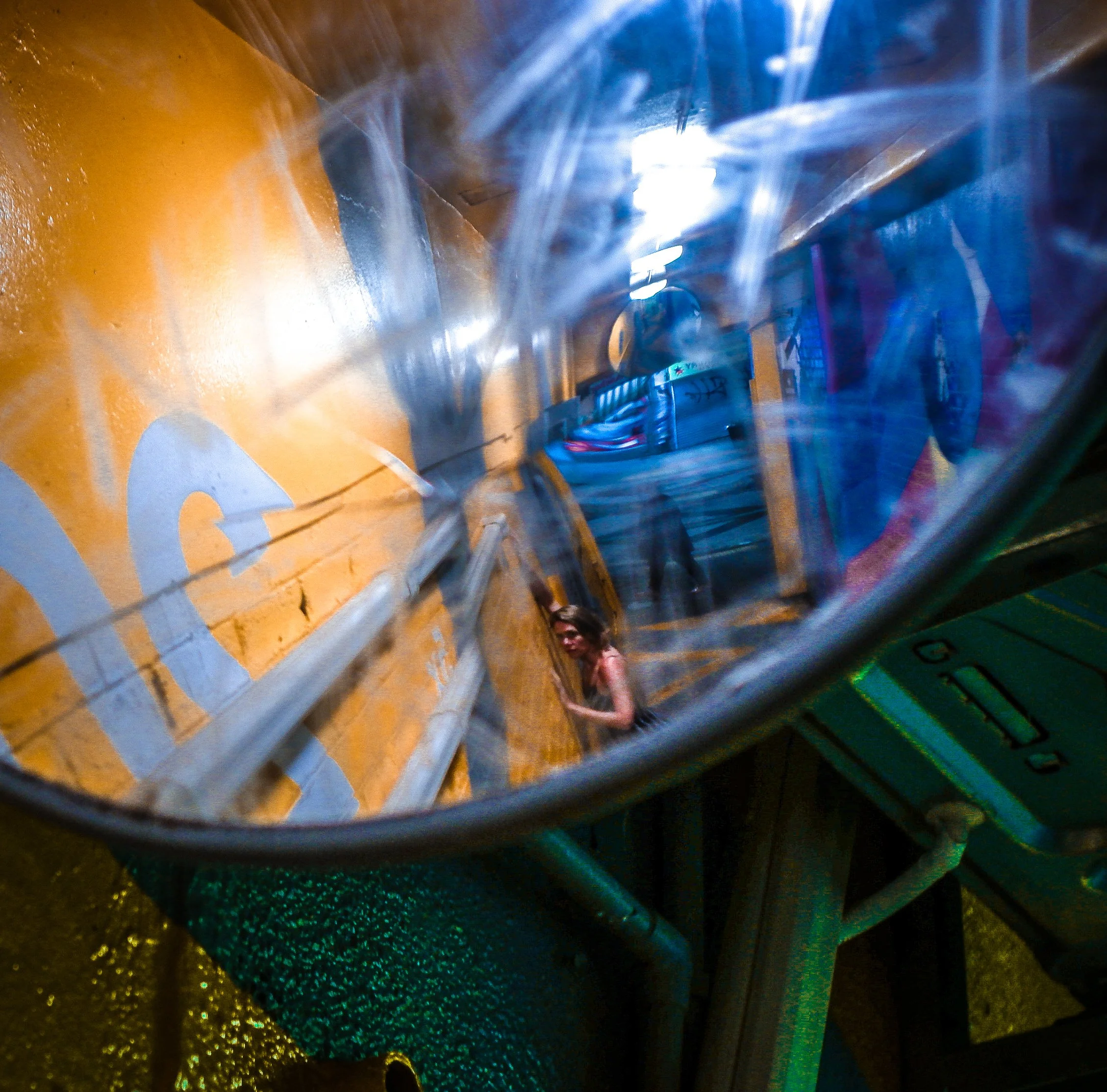 Reflection of a person in a distorted, curved mirror with vibrant blue and yellow surroundings, and machinery or equipment visible in the background.