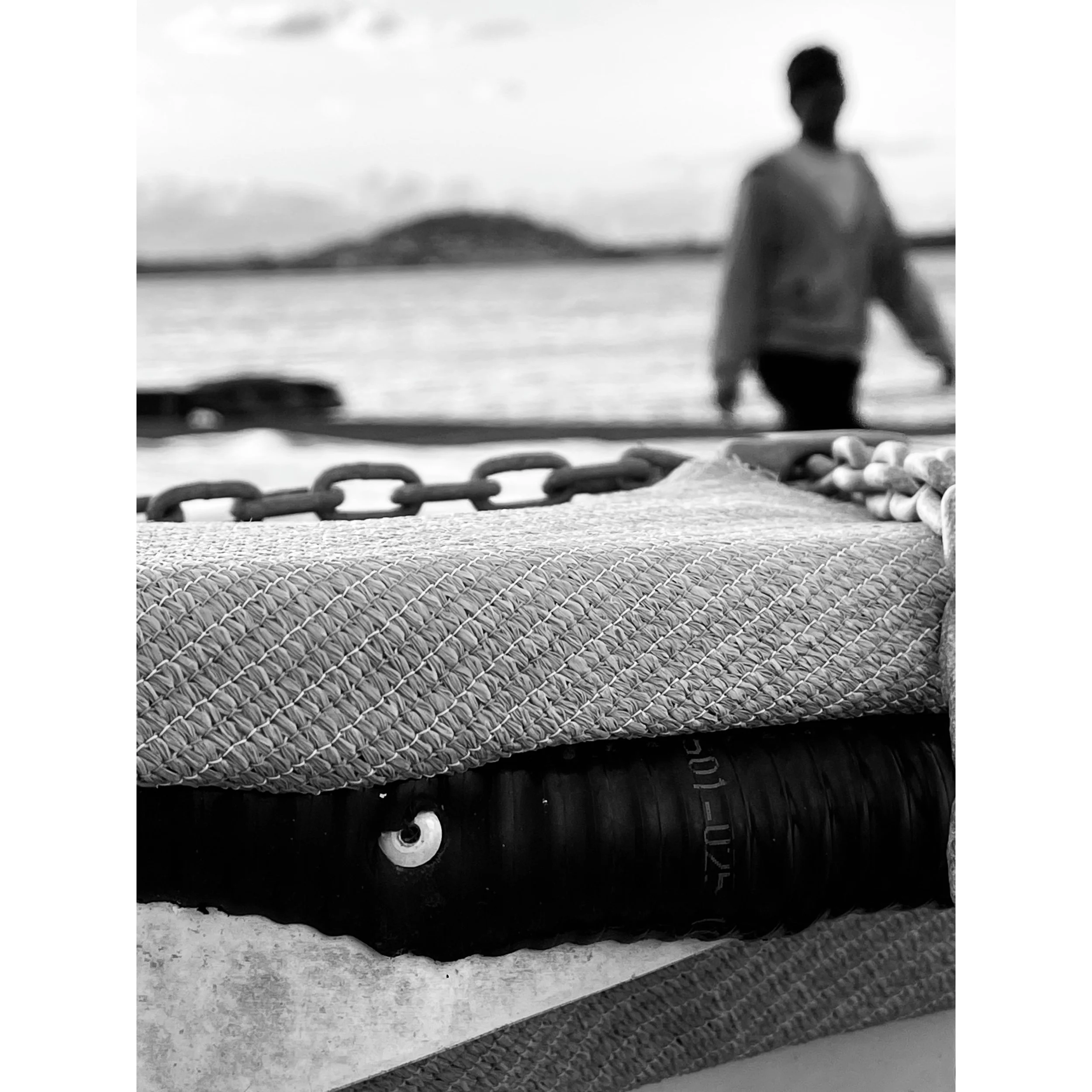 Close-up of a textured surface with a chain and a cylindrical object underneath, with a person walking near water in the background.