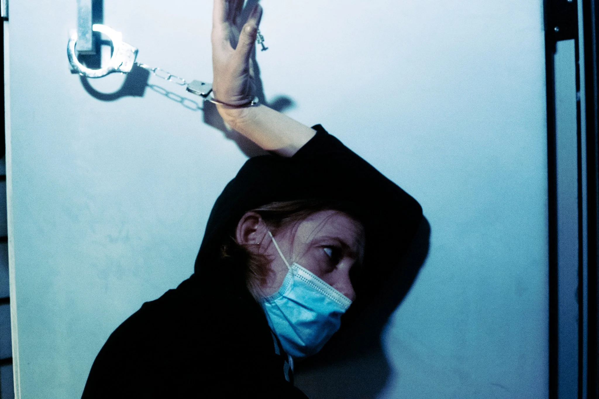 A woman with red hair wearing a face mask, with her head resting against a wall, and an arm raised above her head holding broken handcuffs attached to a chain.