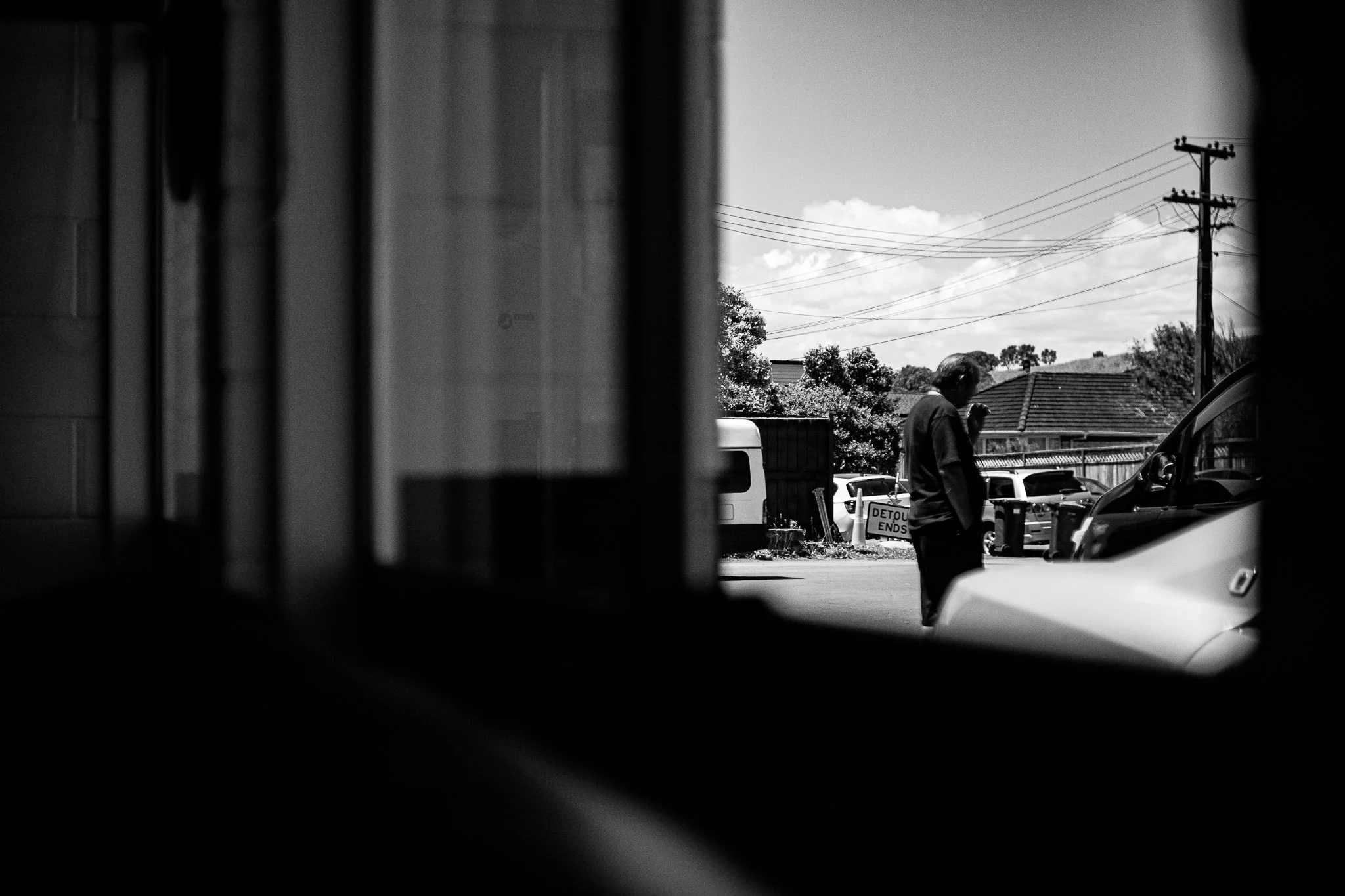 A person stands outside near parked cars, seen through a window in black and white, with power lines and trees in the background.