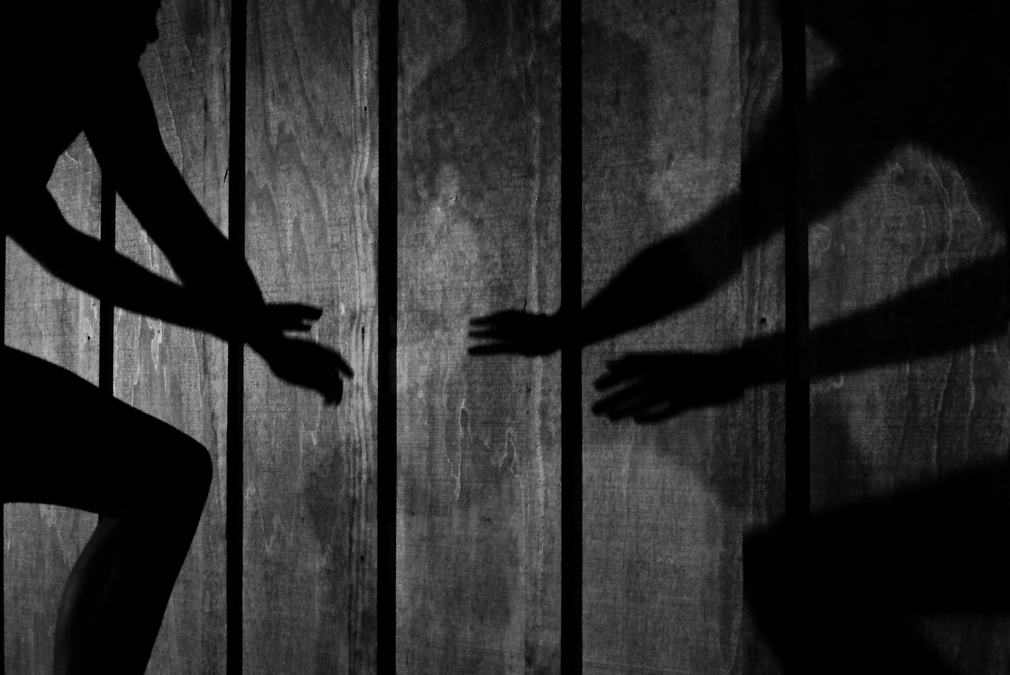 Silhouettes of two hands reaching toward each other behind prison bars in a black and white photograph.