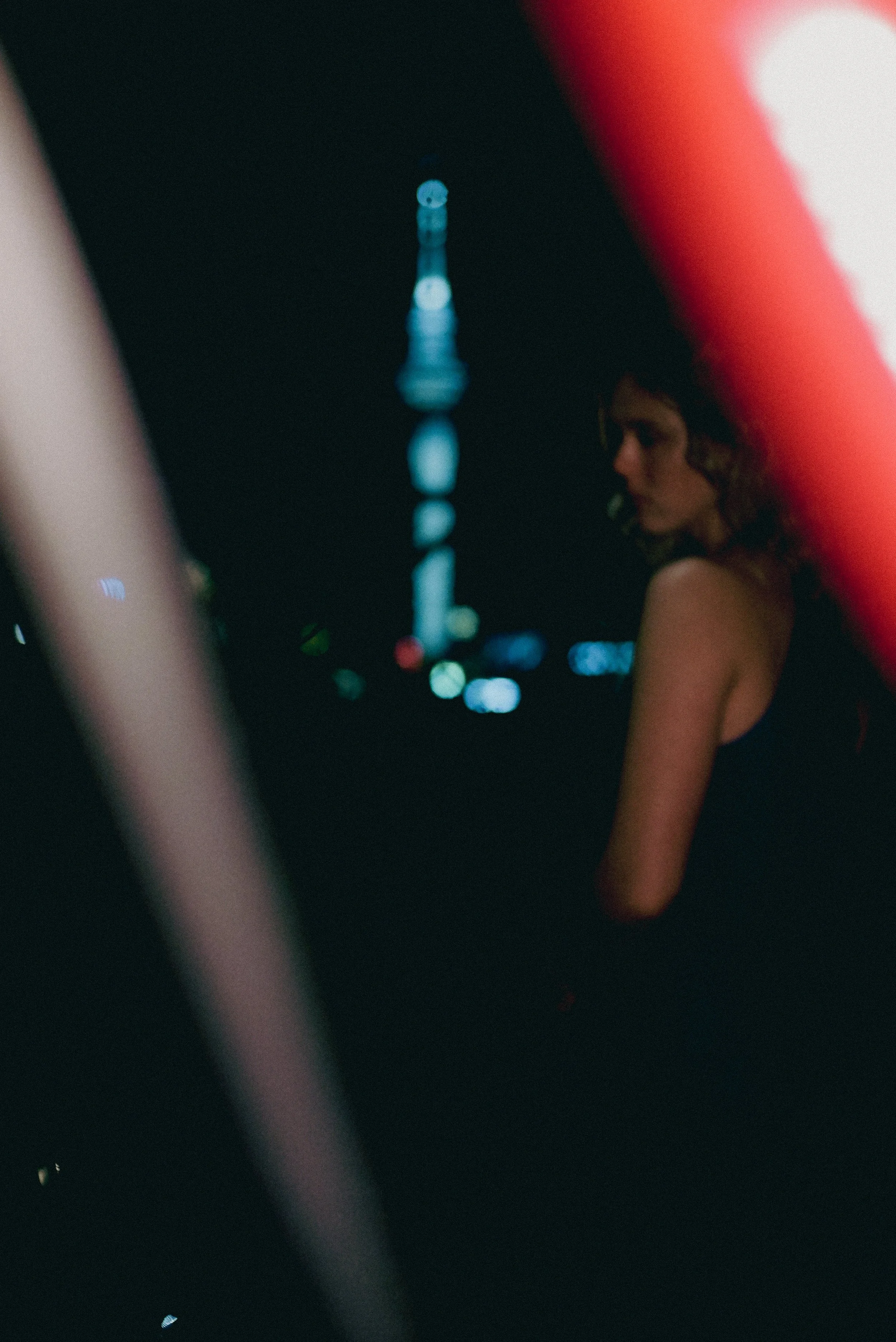 Nighttime photo of a woman with dark curly hair looking to her right, with city lights and a tall illuminated tower in the background.