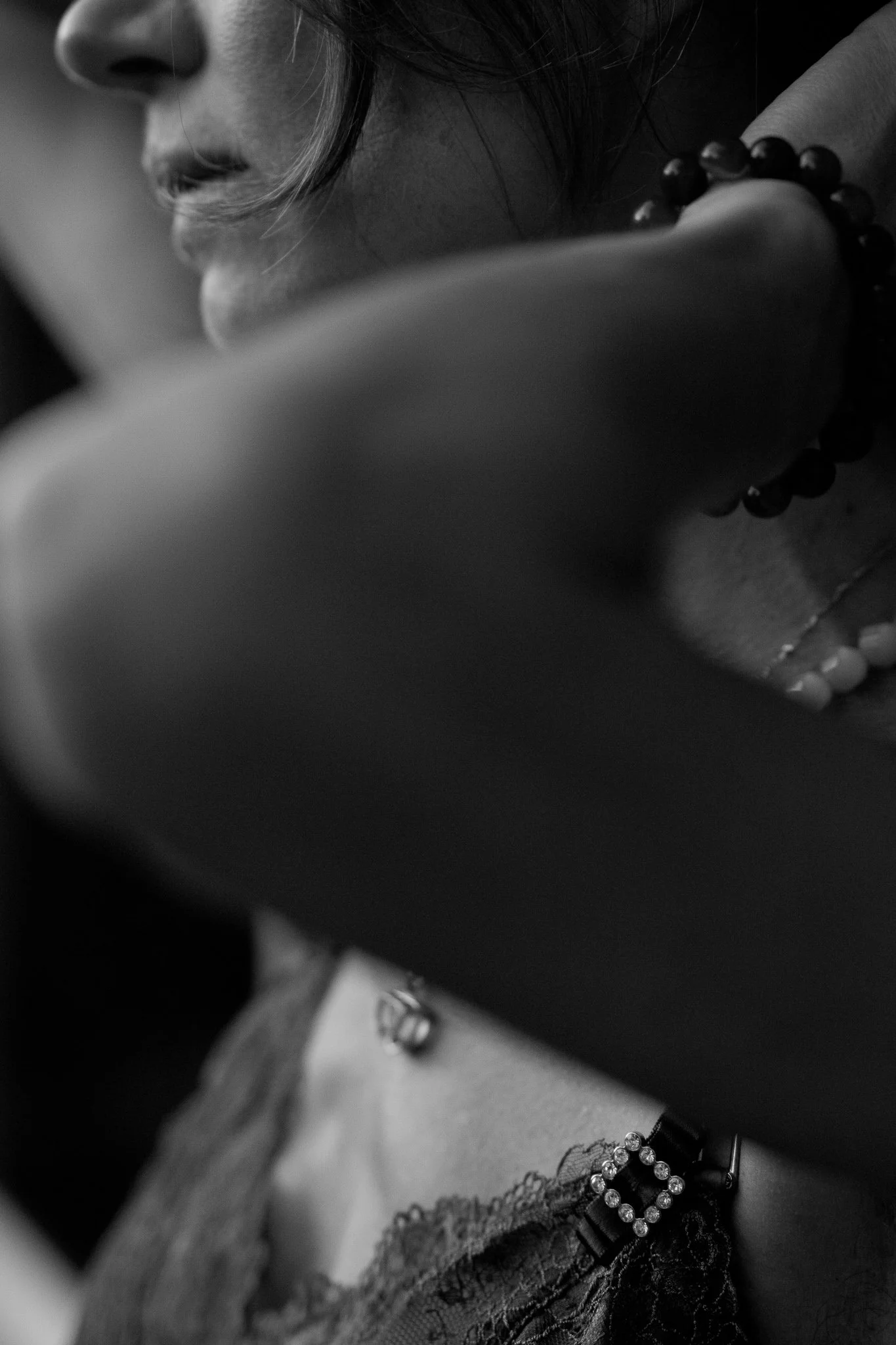 Close-up black and white photo of a woman's face, with her lips slightly parted, wearing a beaded bracelet, and her chin resting on her arm, with some jewelry visible.