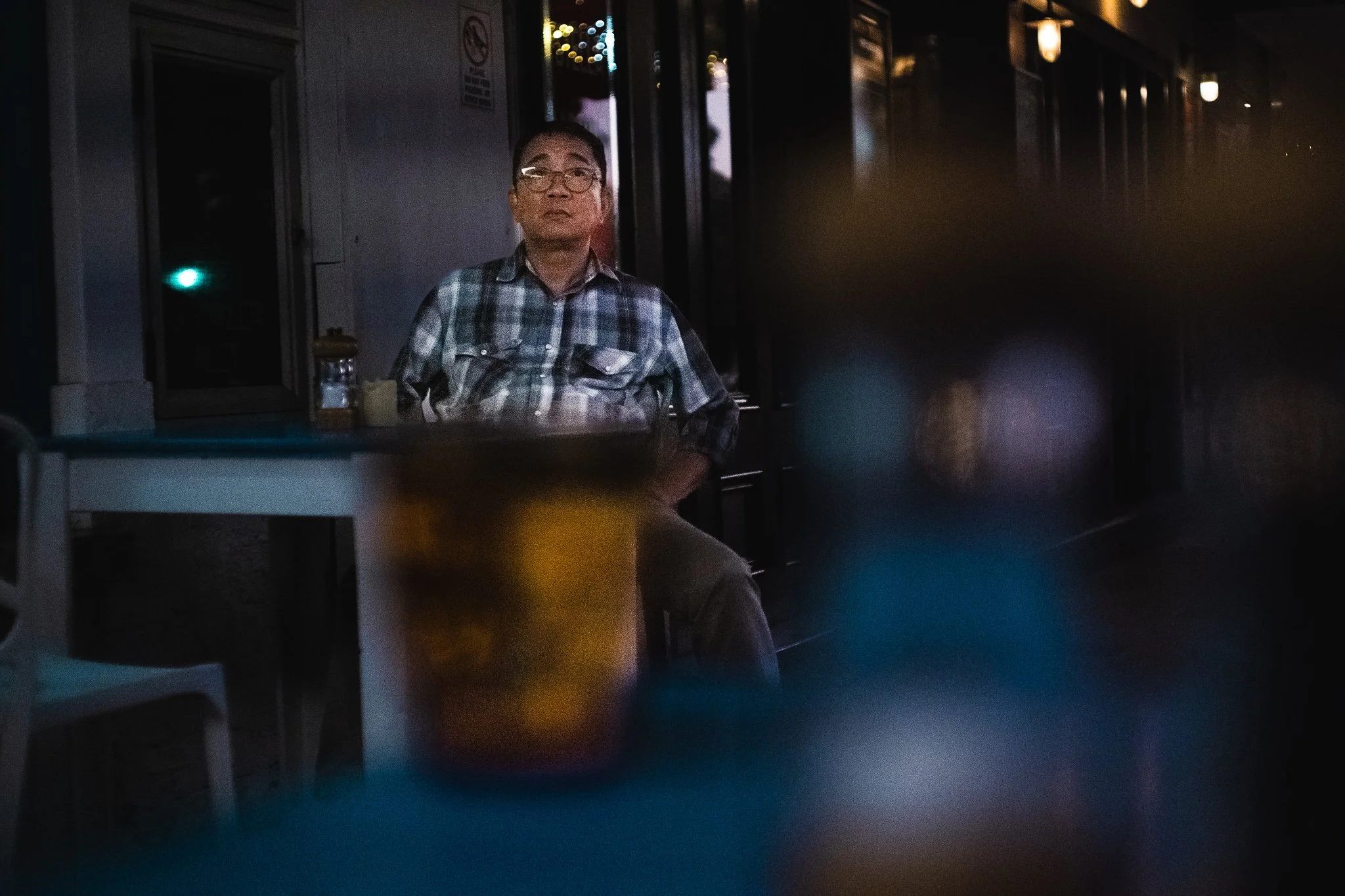 A man wearing glasses and a checkered shirt sits at a table in a dimly lit restaurant or cafe, with a blurred foreground and a string of lights outside the window behind him.