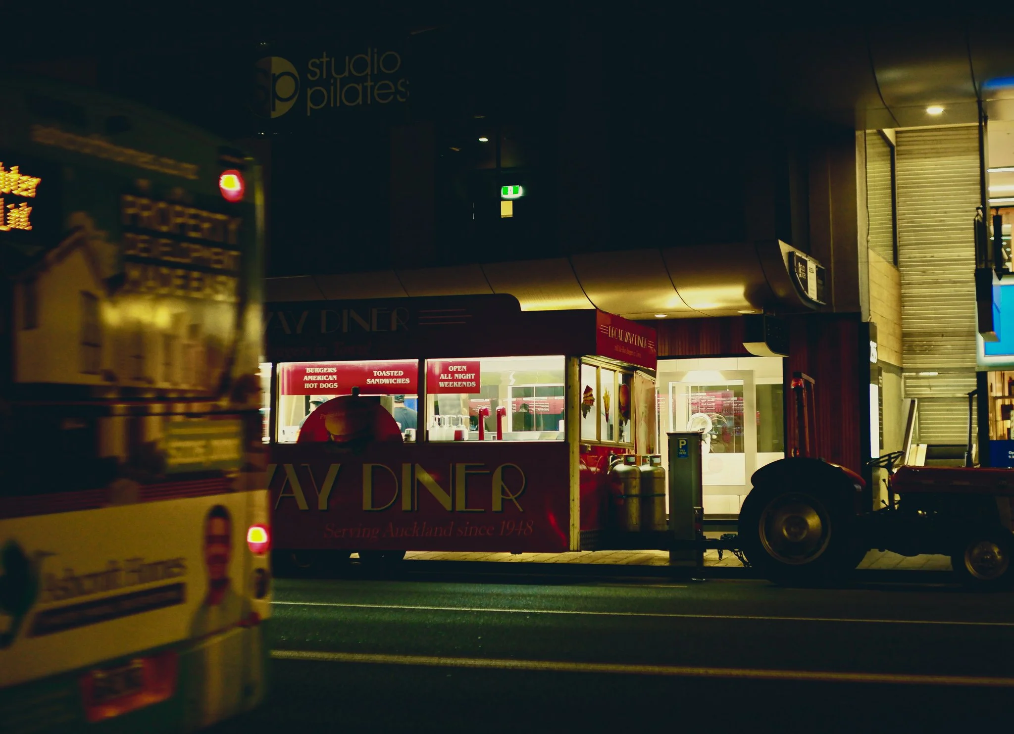 Nighttime scene of a retro-style diner on a city street, with a food truck and a parked tractor, illuminated by warm lighting, and signage indicating it's a 24-hour eatery serving burgers, American hot dogs, and toasted sandwiches.