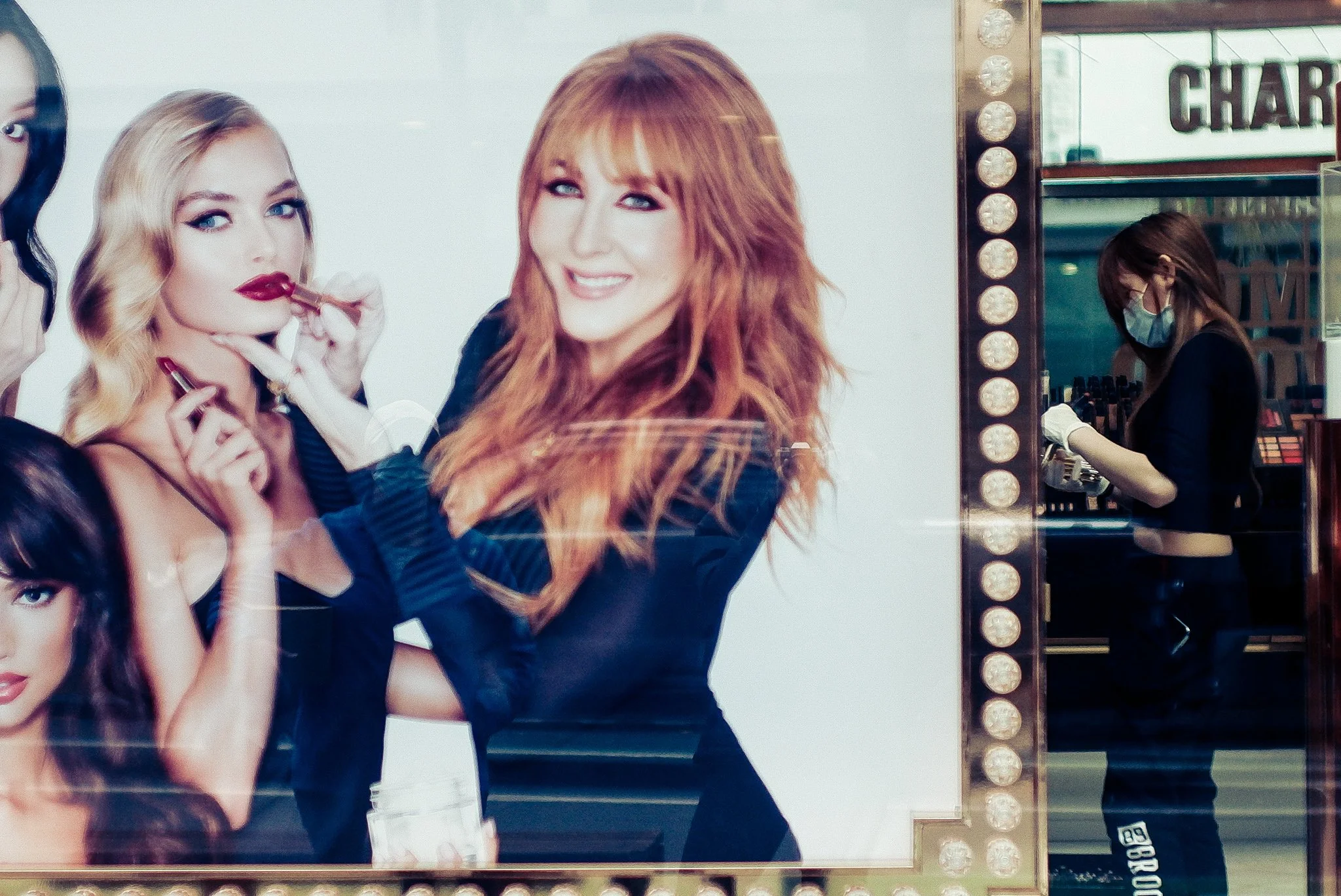 A store display features a large image of four women, three with blonde or red hair, one with dark hair, applying lipstick. A woman wearing a mask and gloves is seen in the background, working with cosmetics inside the store.