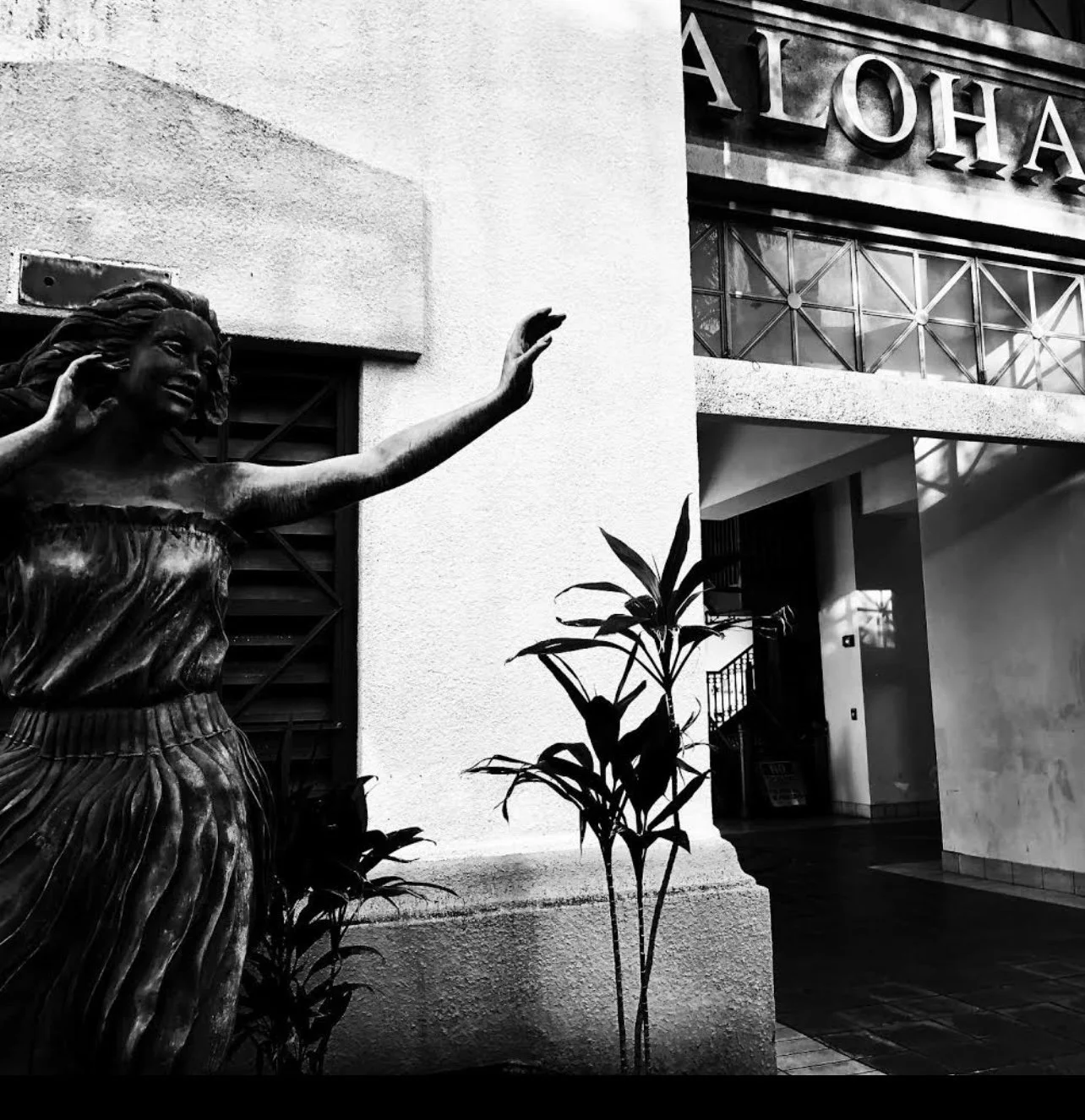 Black and white photo of a bronze statue of a woman with long hair, smiling, with one arm raised, outside a building with the word 'ALOHA' on a sign.