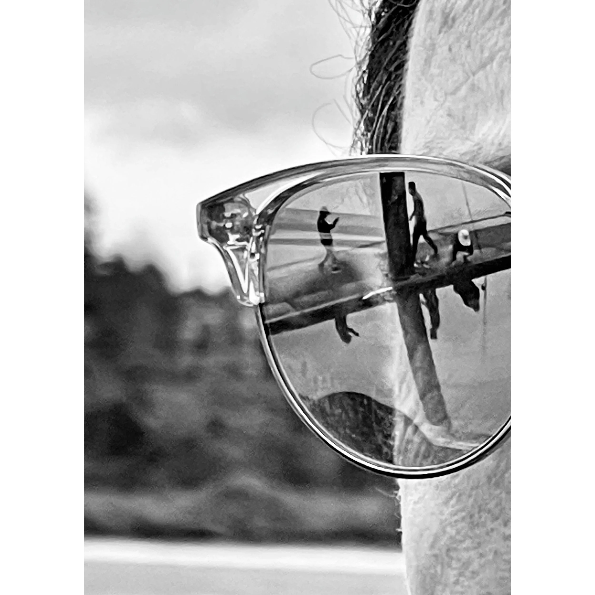 Close-up of sunglasses with reflection of two people walking outdoors on a cloudy day, with trees and sky in the background.