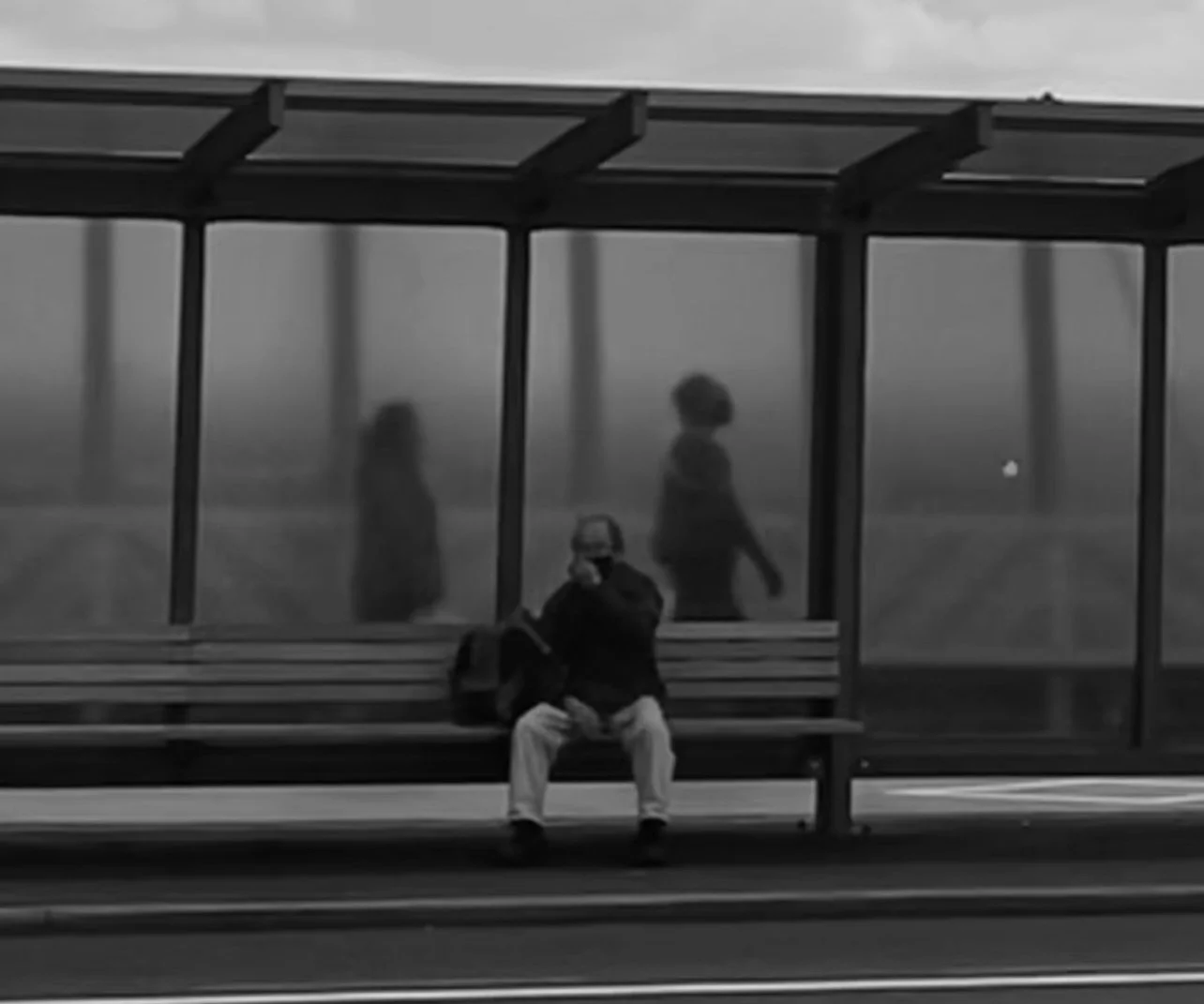 A man sitting on a bench at a foggy bus stop shelter with two women walking in the background.
