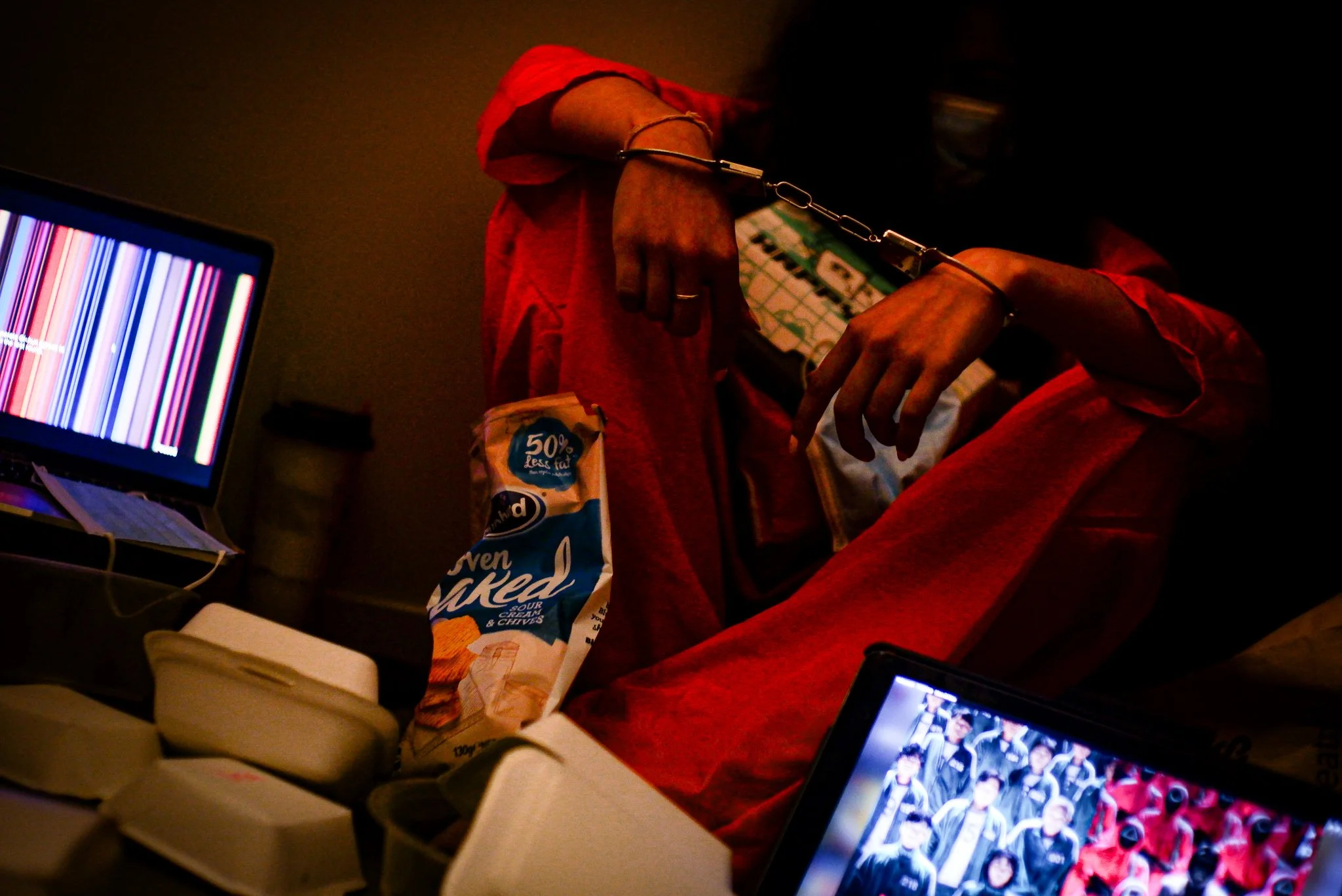 Person wearing a red jacket and red pants, with hands cuffed with metal handcuffs, sitting at a cluttered desk with laptops and snacks in a dark room.