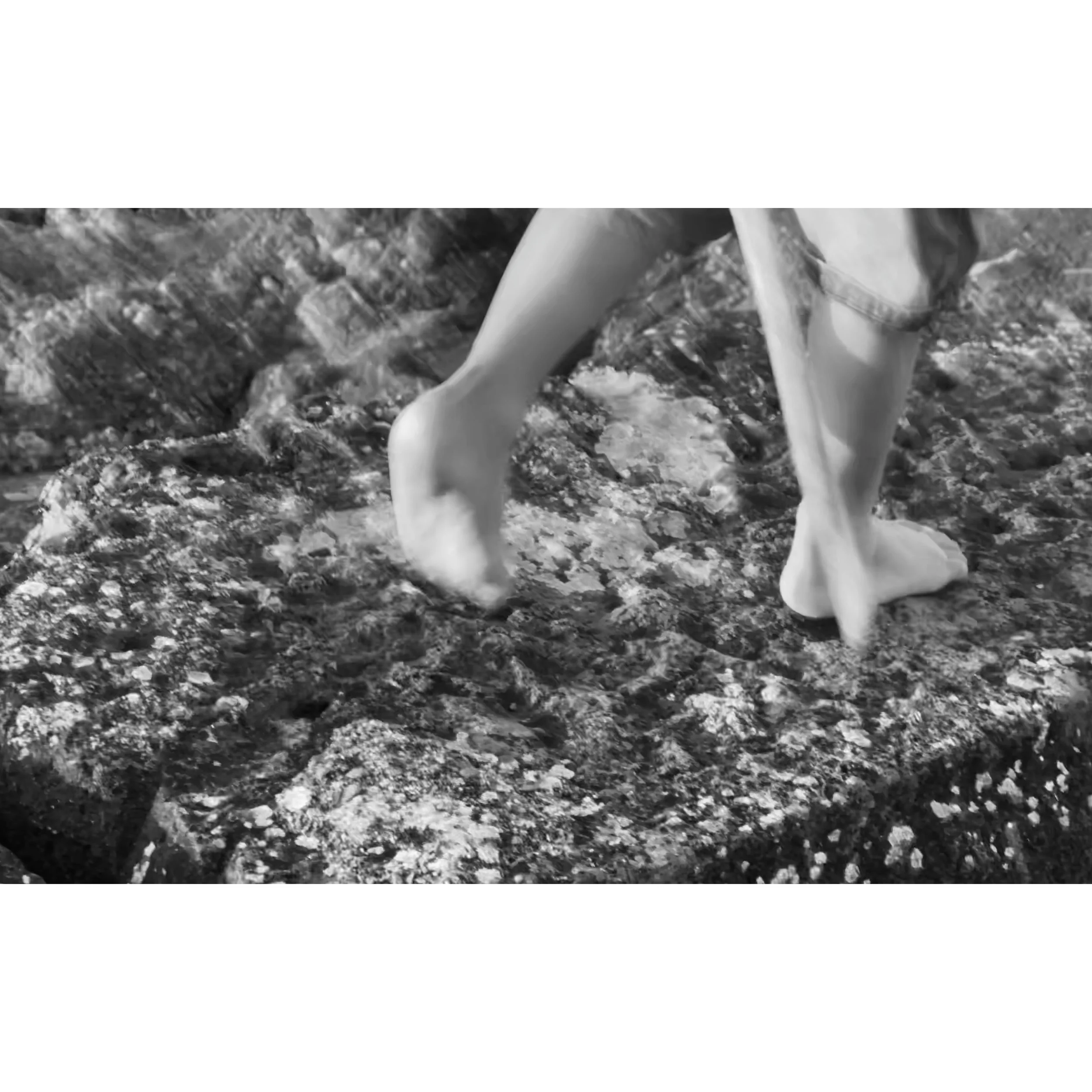 Close-up of bare feet walking on a rocky surface, wearing rolled-up pants, in black and white.