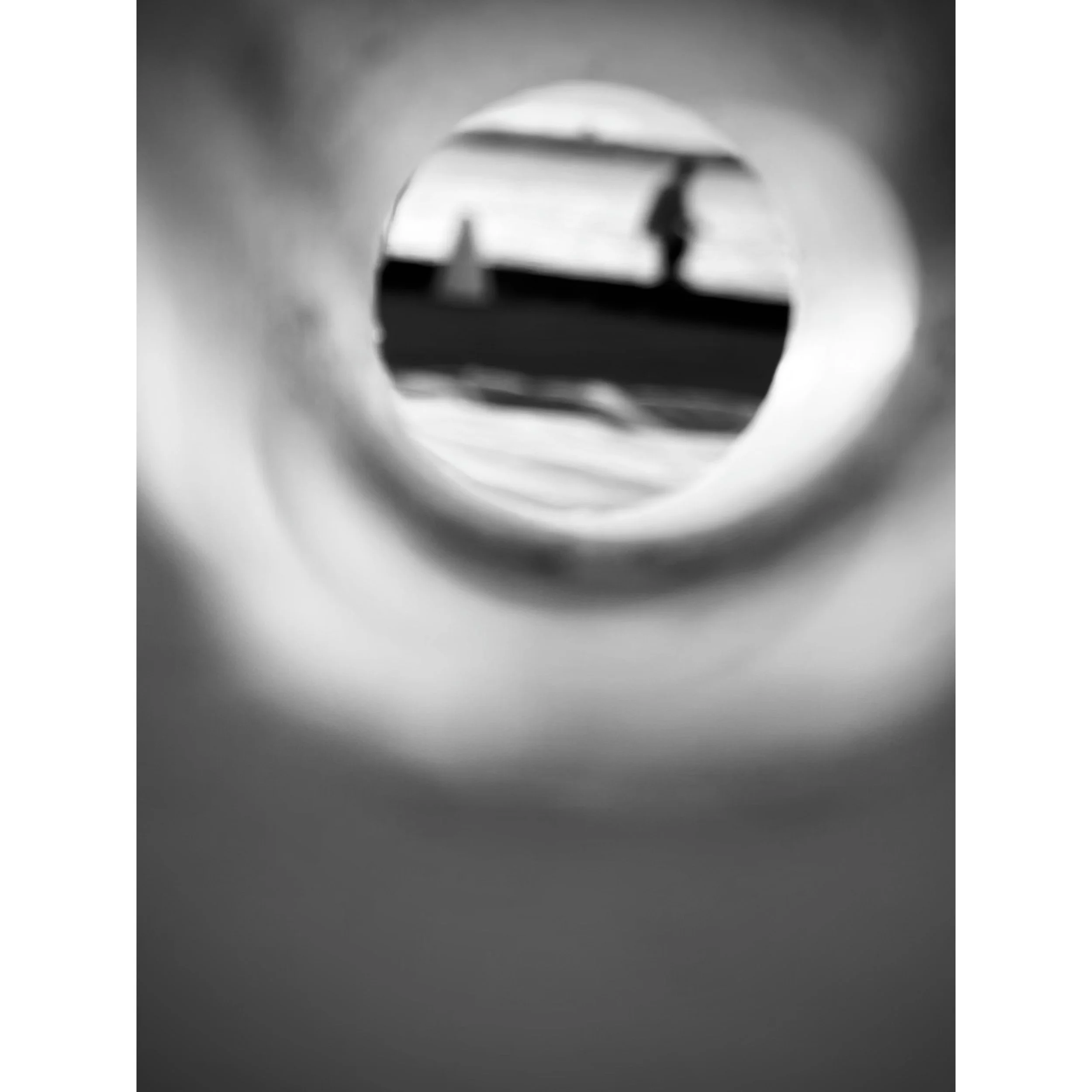 Black and white photograph of a person walking under an umbrella, seen through a circular opening, with a blurred background.