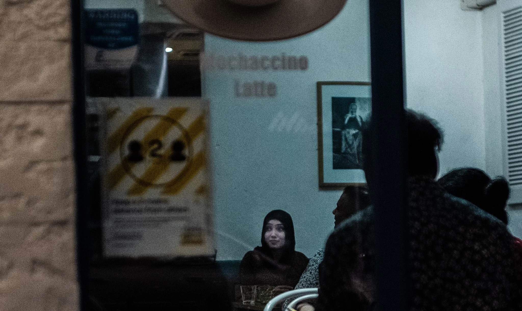 People sitting at a table in a dimly lit restaurant, with a framed photo on the wall and a person in a dark hoodie speaking to others.
