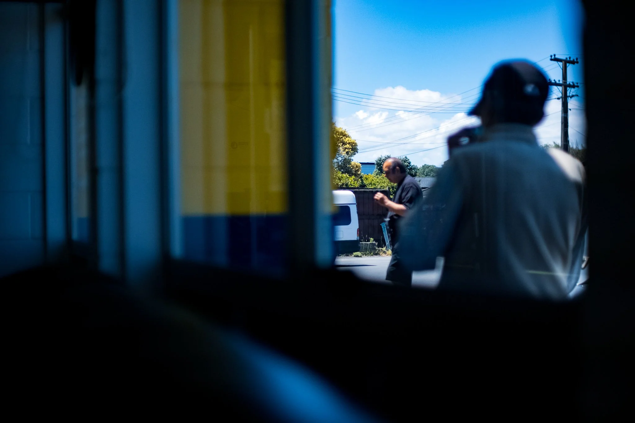 View of two men standing outside, one wearing a dark shirt looking at their phone and the other in a light-colored jacket and cap, seen through a small window or opening.