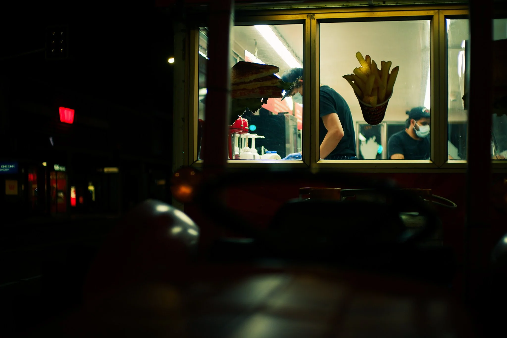 View through a window showing three people working behind a food truck counter, one wearing a sandwich hat and sneakers, all wearing face masks, with french fries in a cone and a sandwich on the window.
