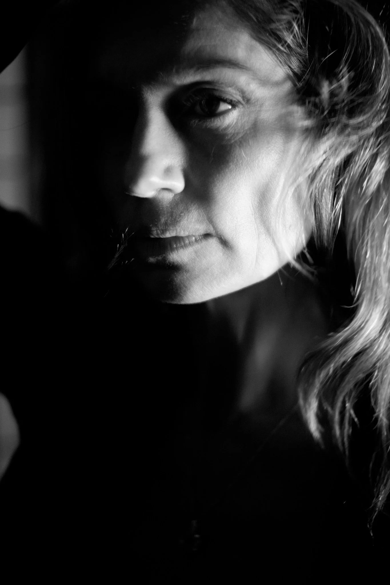 A black and white close-up portrait of a woman with wavy hair, looking to the side, with dramatic lighting casting deep shadows on one side of her face.