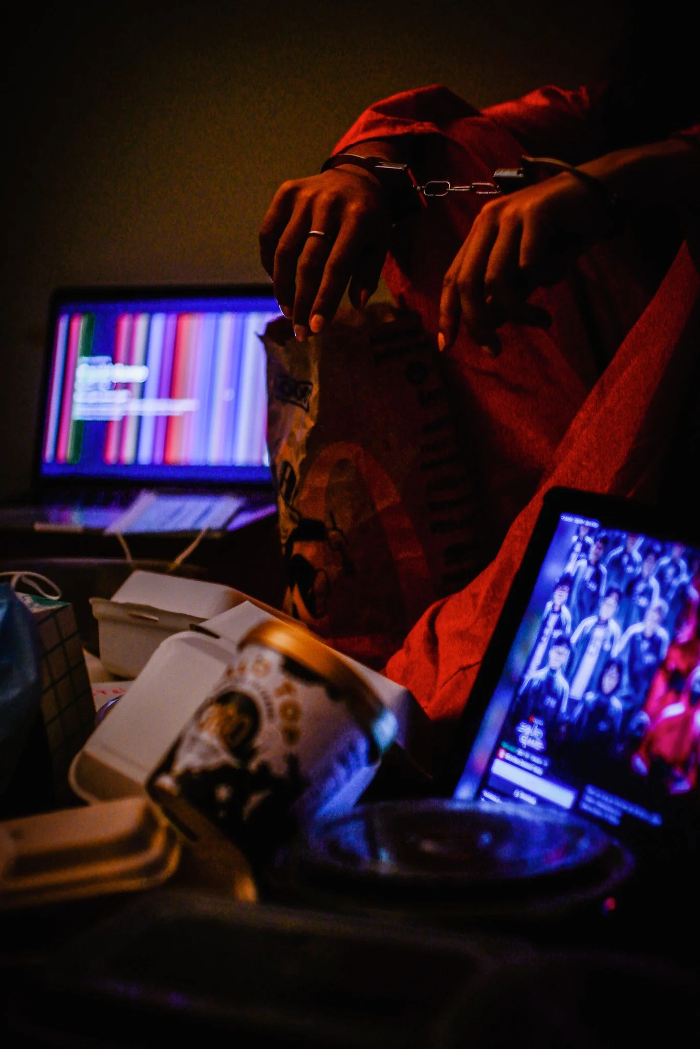 A person wearing an orange jacket and handcuffs, sitting in a dark room with a laptop, tablet, and snacks, watching a sports event on the devices.