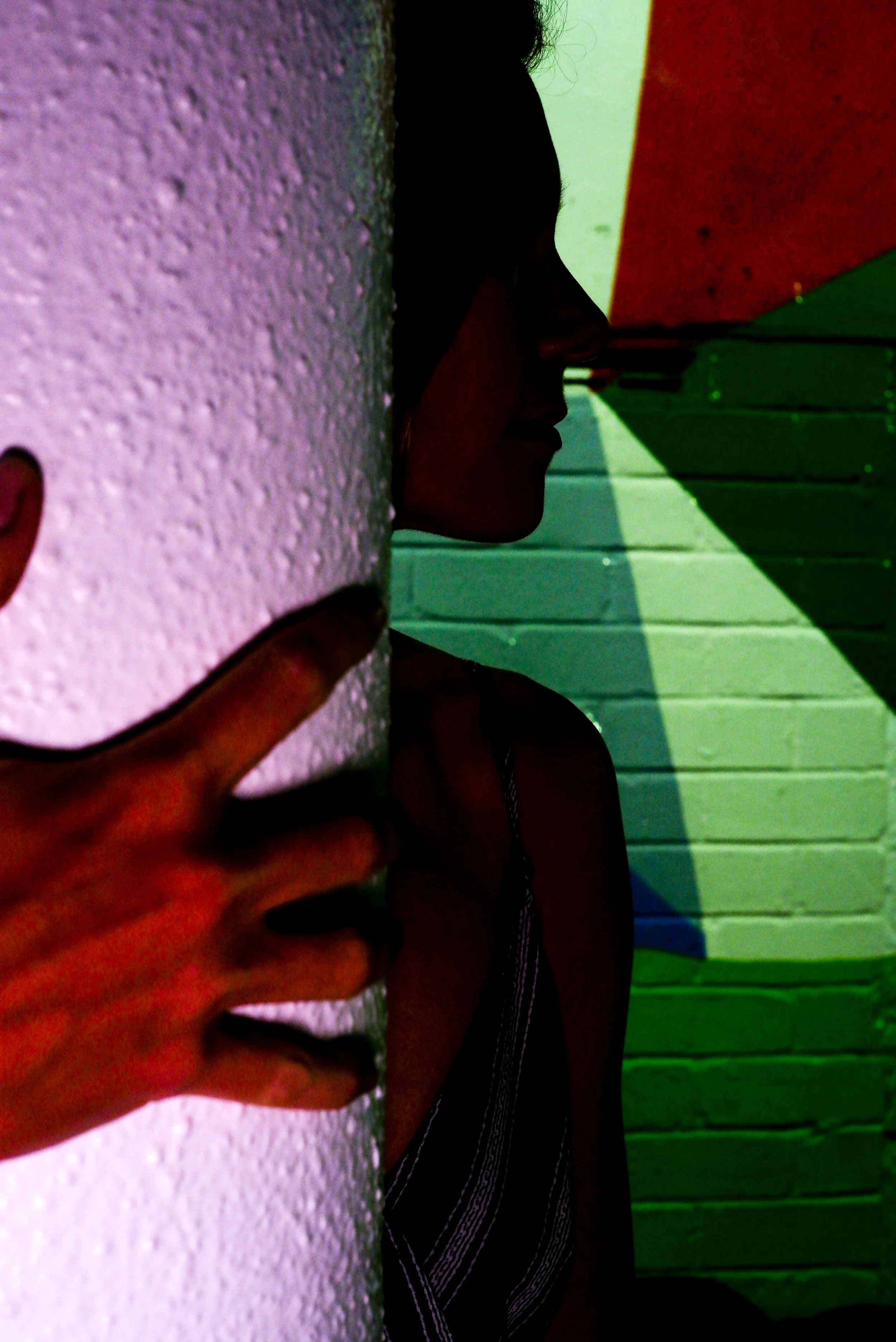 A person making a peace sign with their hand while standing against a textured wall, with part of their face and body partially obscured by the wall. The background features a colorful, geometric wall design.