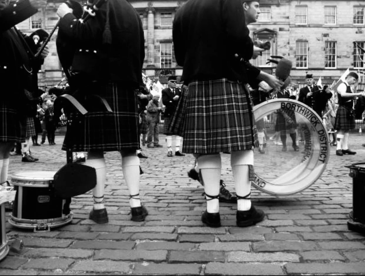 People in traditional Scottish attire, including kilts, playing musical instruments in a street setting.