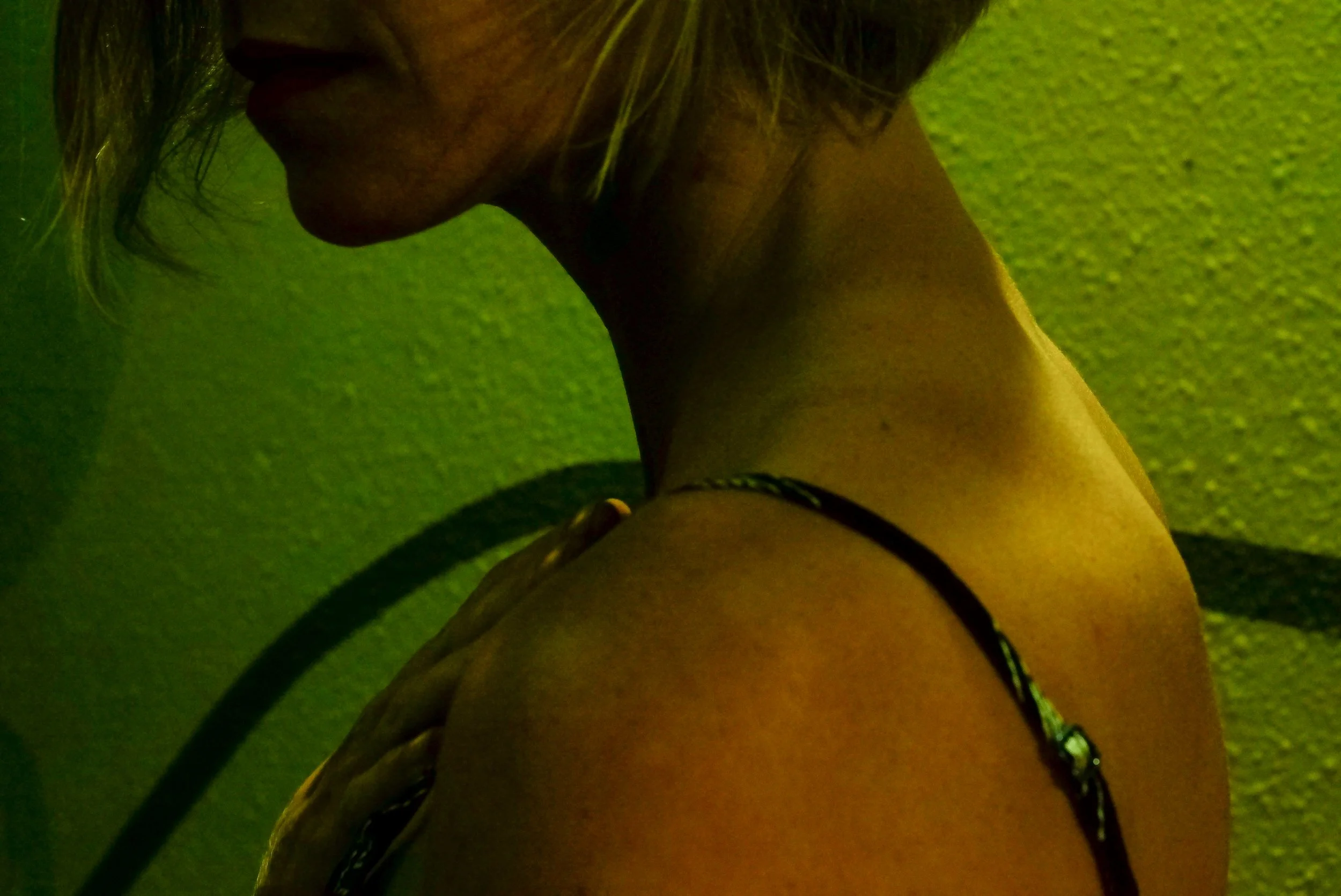 A close-up side profile of a person's neck and shoulder, with blonde hair, against a green textured wall.