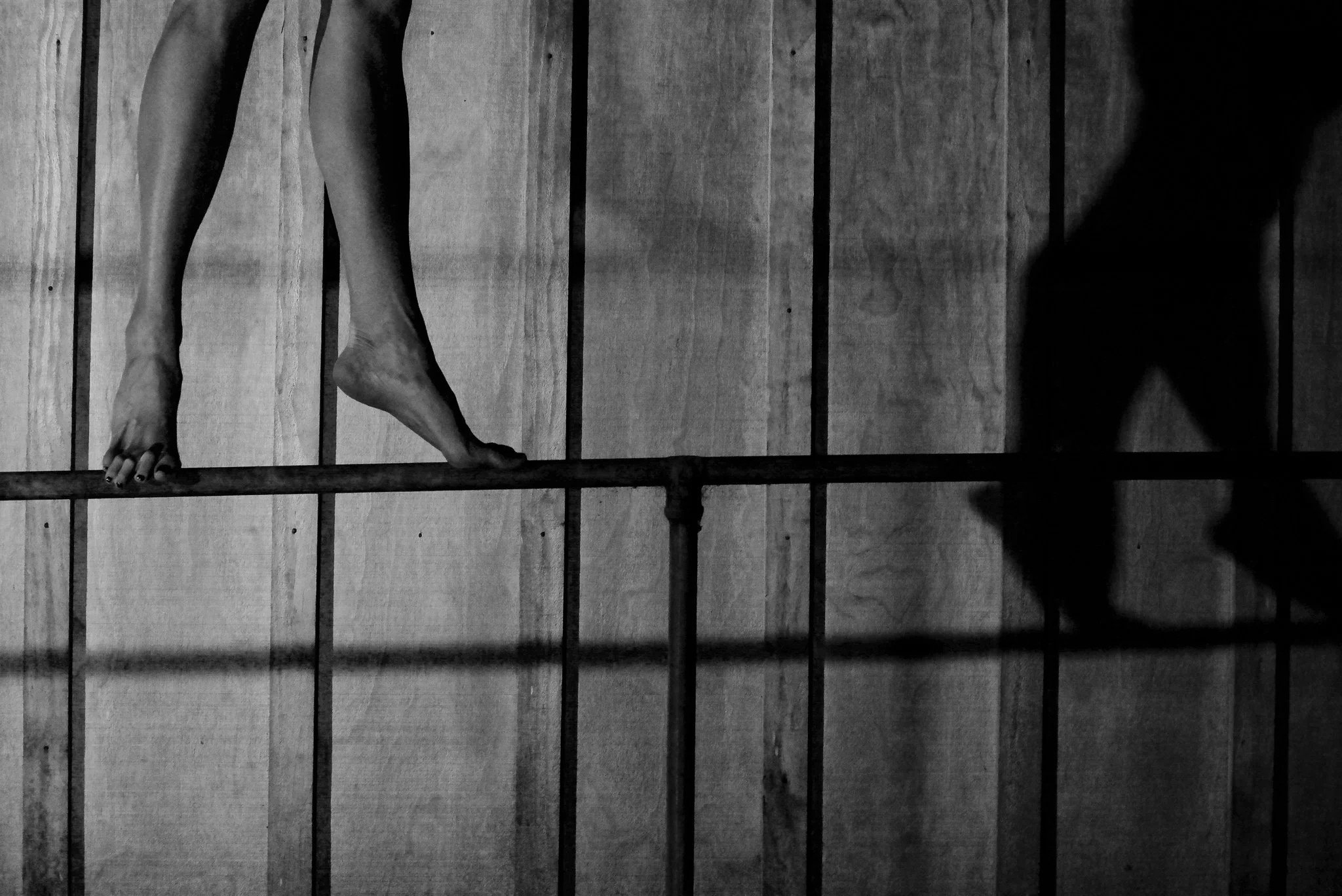 A pair of human legs and feet are visible, with one foot on a horizontal bar and another foot on the ground. The background consists of wooden vertical panels, and there is a prominent shadow cast on the panels. The photo is in black and white.