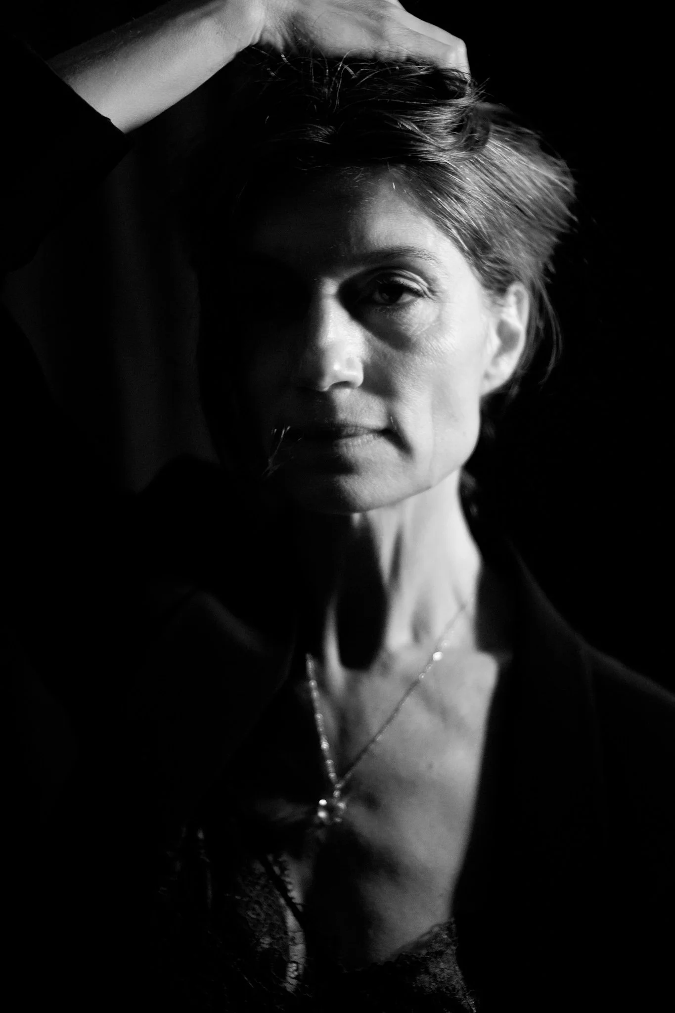 A black and white portrait of a woman with short hair, one hand resting on her head, wearing a necklace and a dark top, looking at the camera.