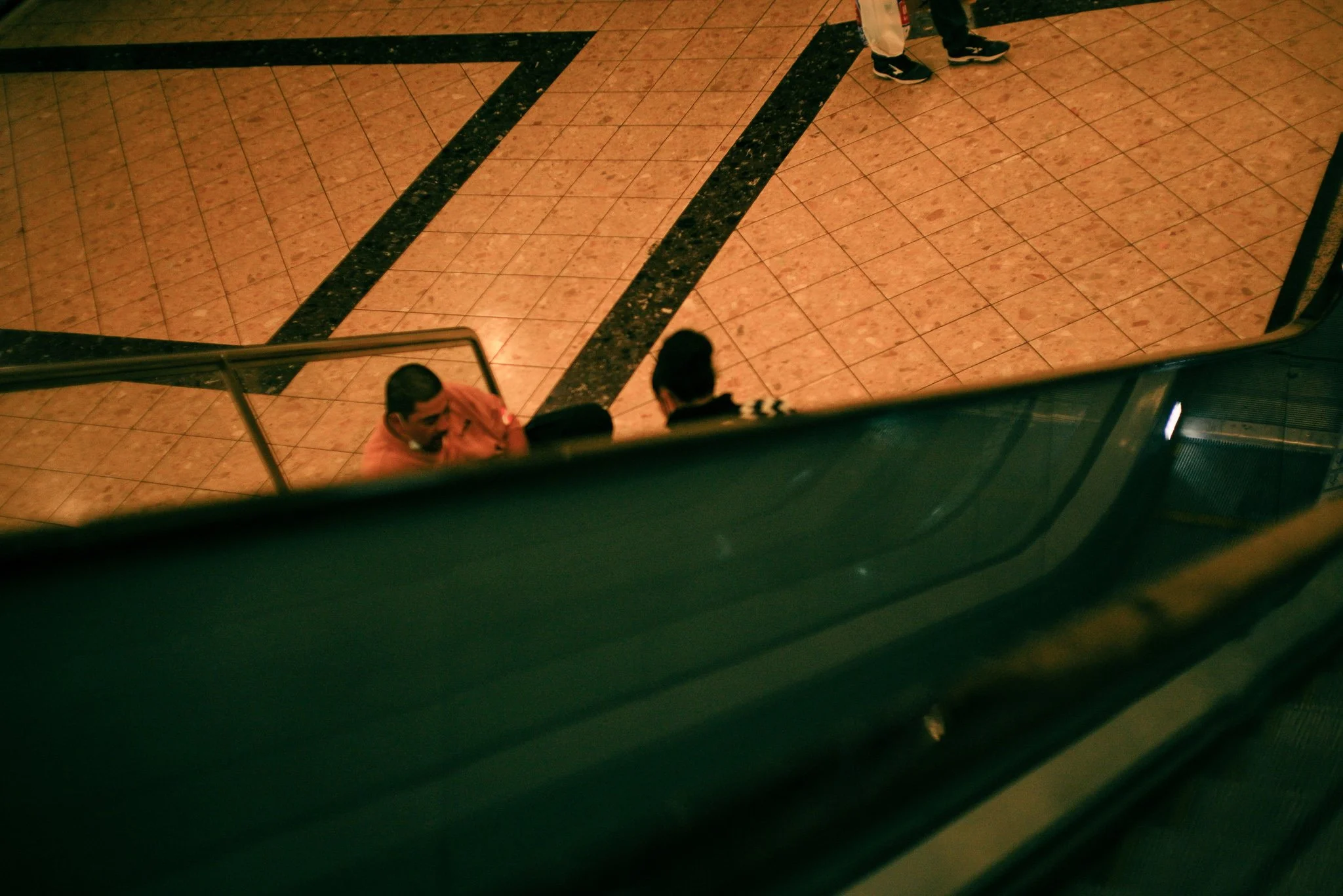 View from an escalator showing two people standing at the bottom, and one person walking down the stairs, inside a building with tiled flooring.