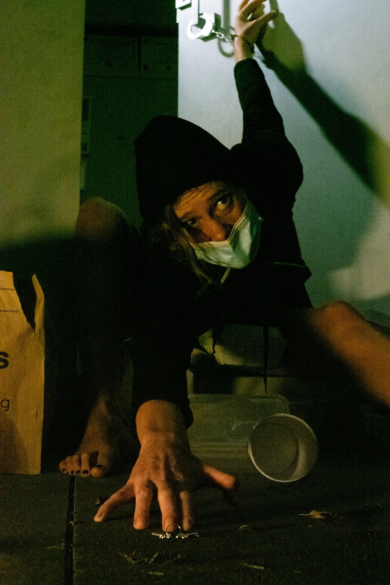 A person wearing a black hoodie and a face mask, reaching out on the floor of a dimly lit room.